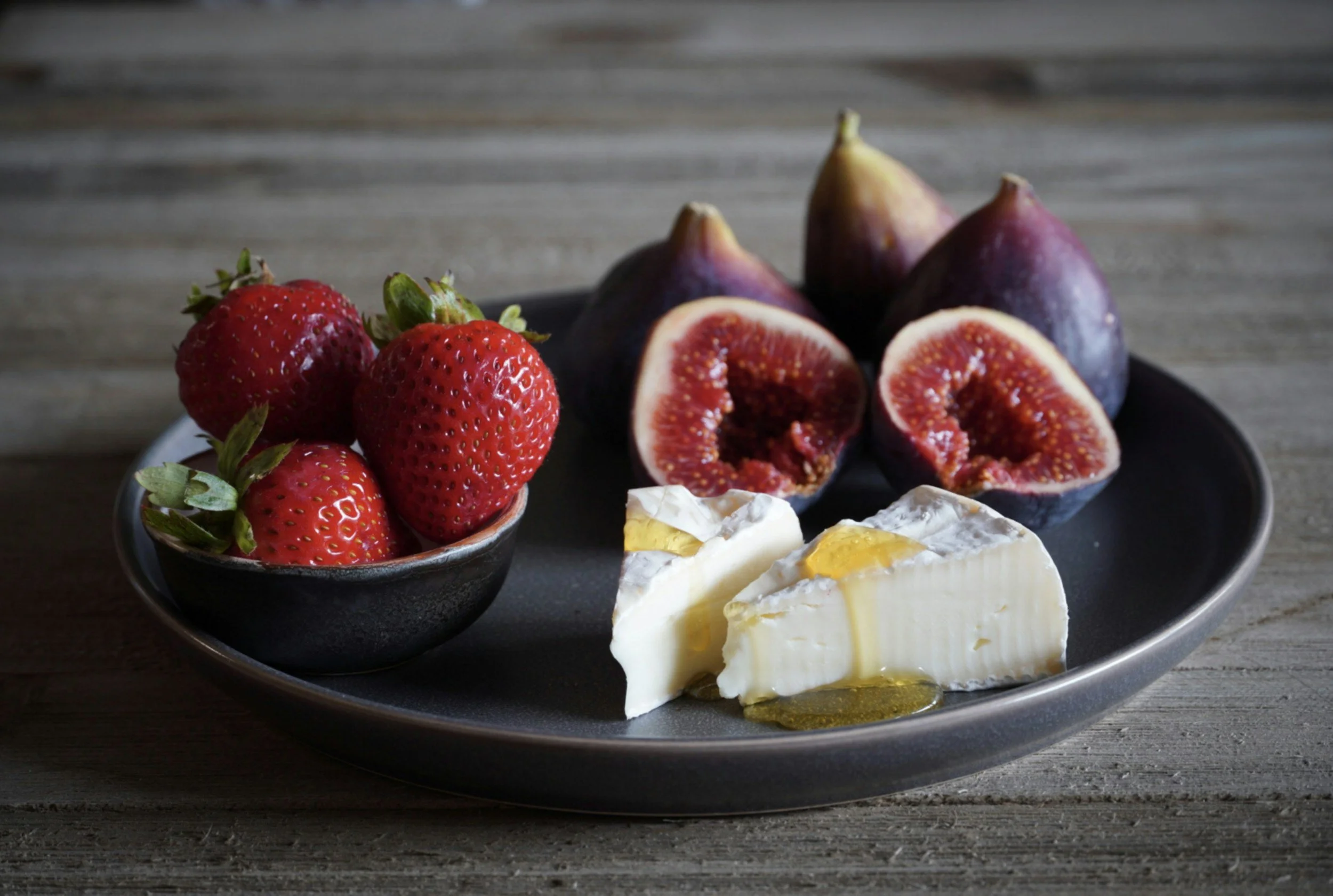 A gray plate with strawberries, figs, and Brie cheese on a wooden surface.