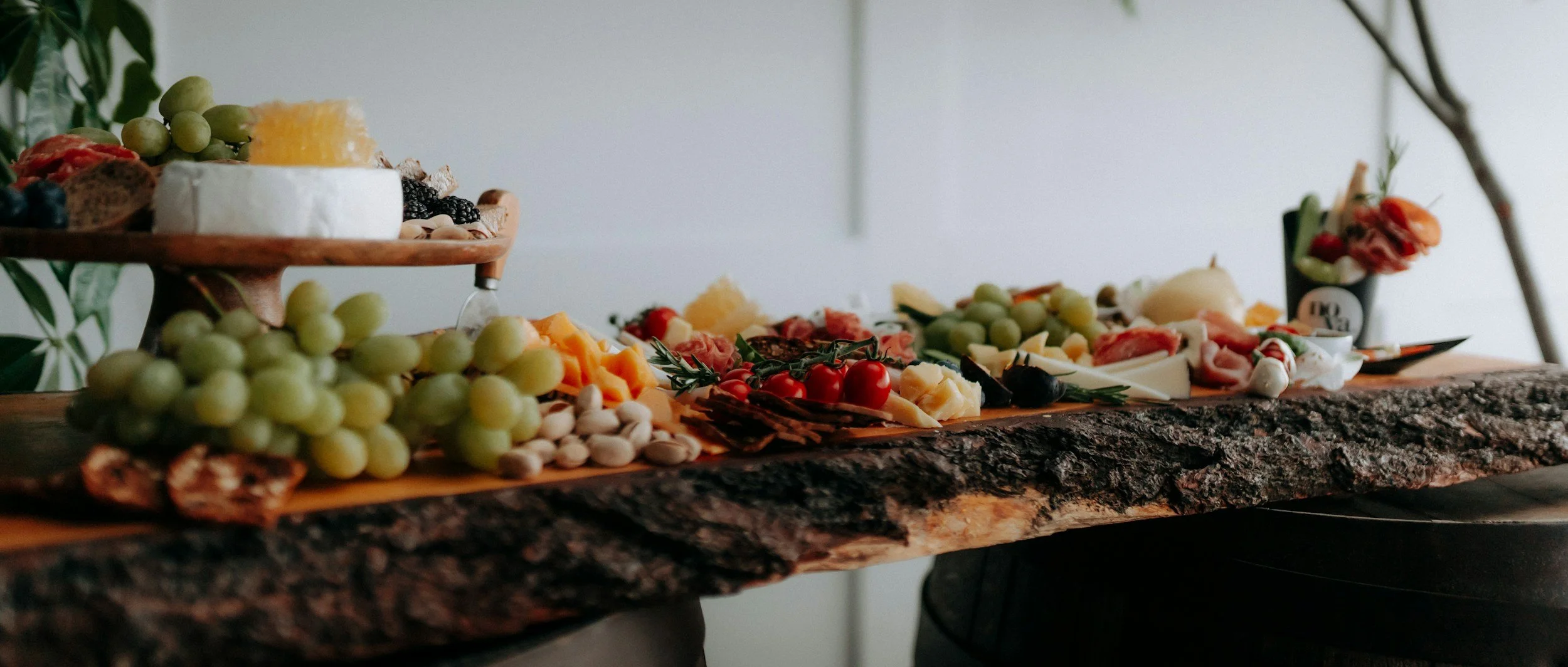 Cheese, grapes, cherry tomatoes, olives, and assorted charcuterie on a rustic wooden serving board and tiered tray, with a small flower arrangement in the background.