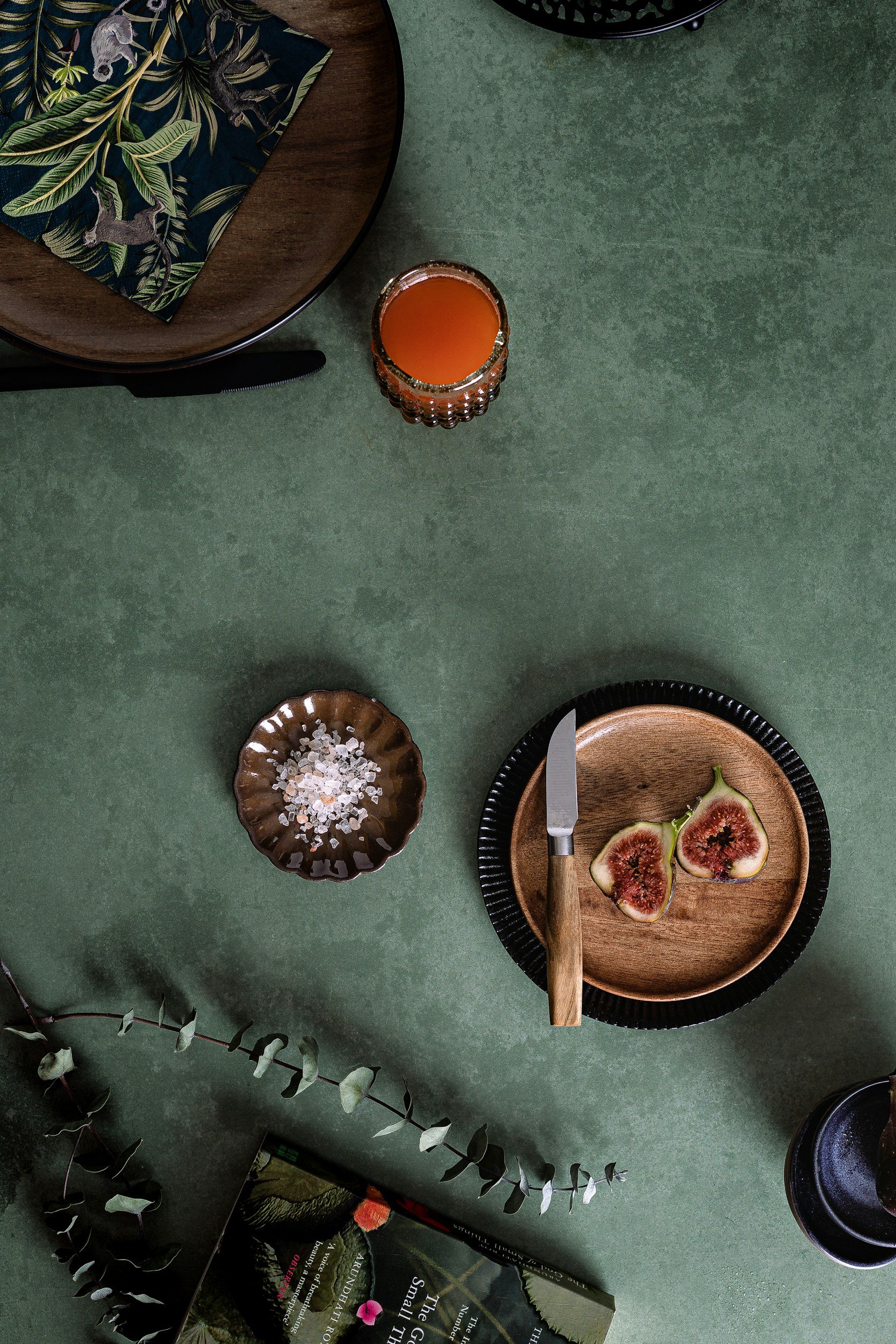 An overhead view of a table setting with a glass of orange juice, a small bowl of coarse salt, a plate with halved figs, a knife, a black-rimmed plate, and a decorative plate with a dark pattern featuring various animals, all on a green table. There are also some books and greenery nearby.