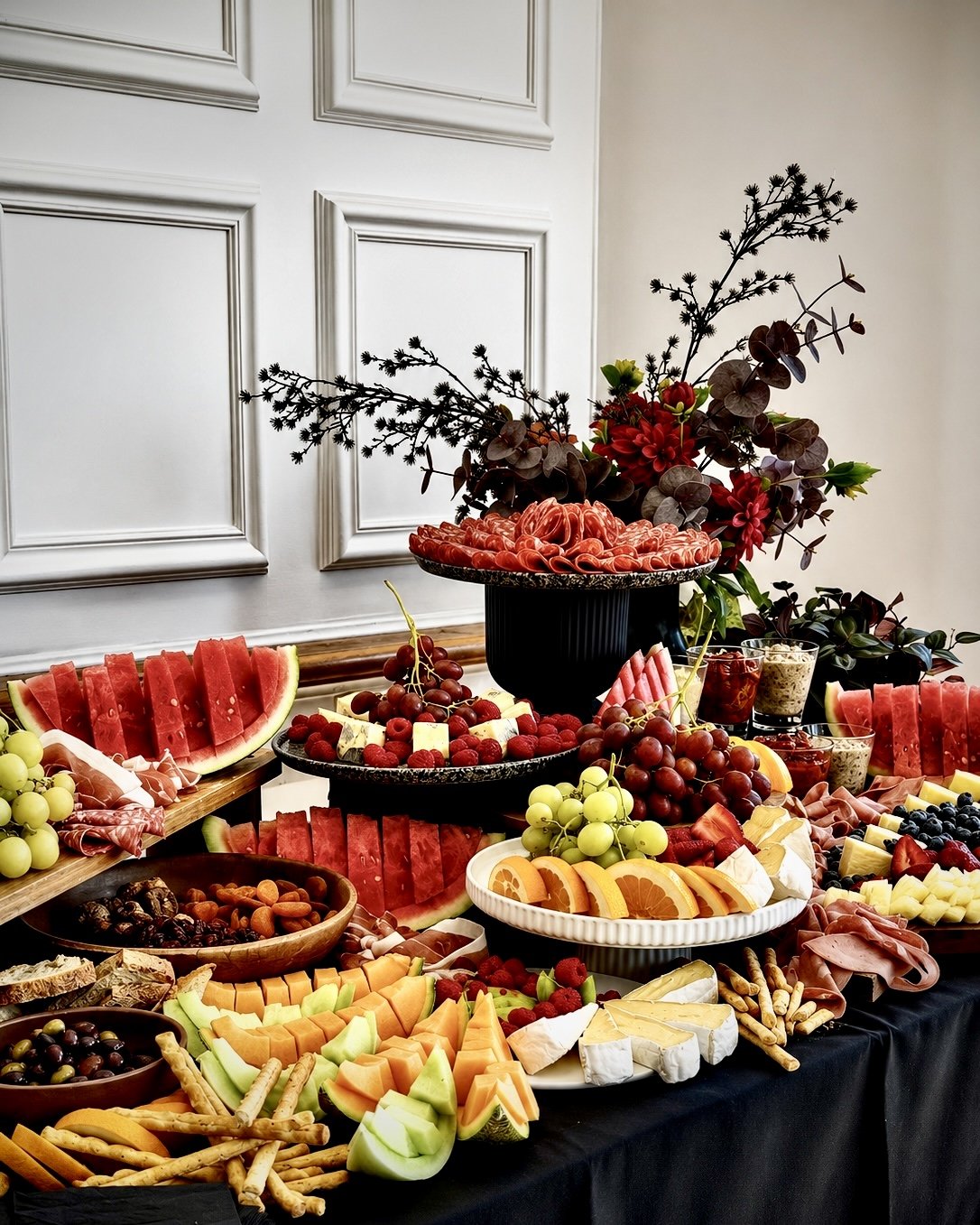 Colorful fruit platter with watermelon, cantaloupe, honeydew, grapes, berries, cheese, charcuterie, and garnished with greenery on a black table.