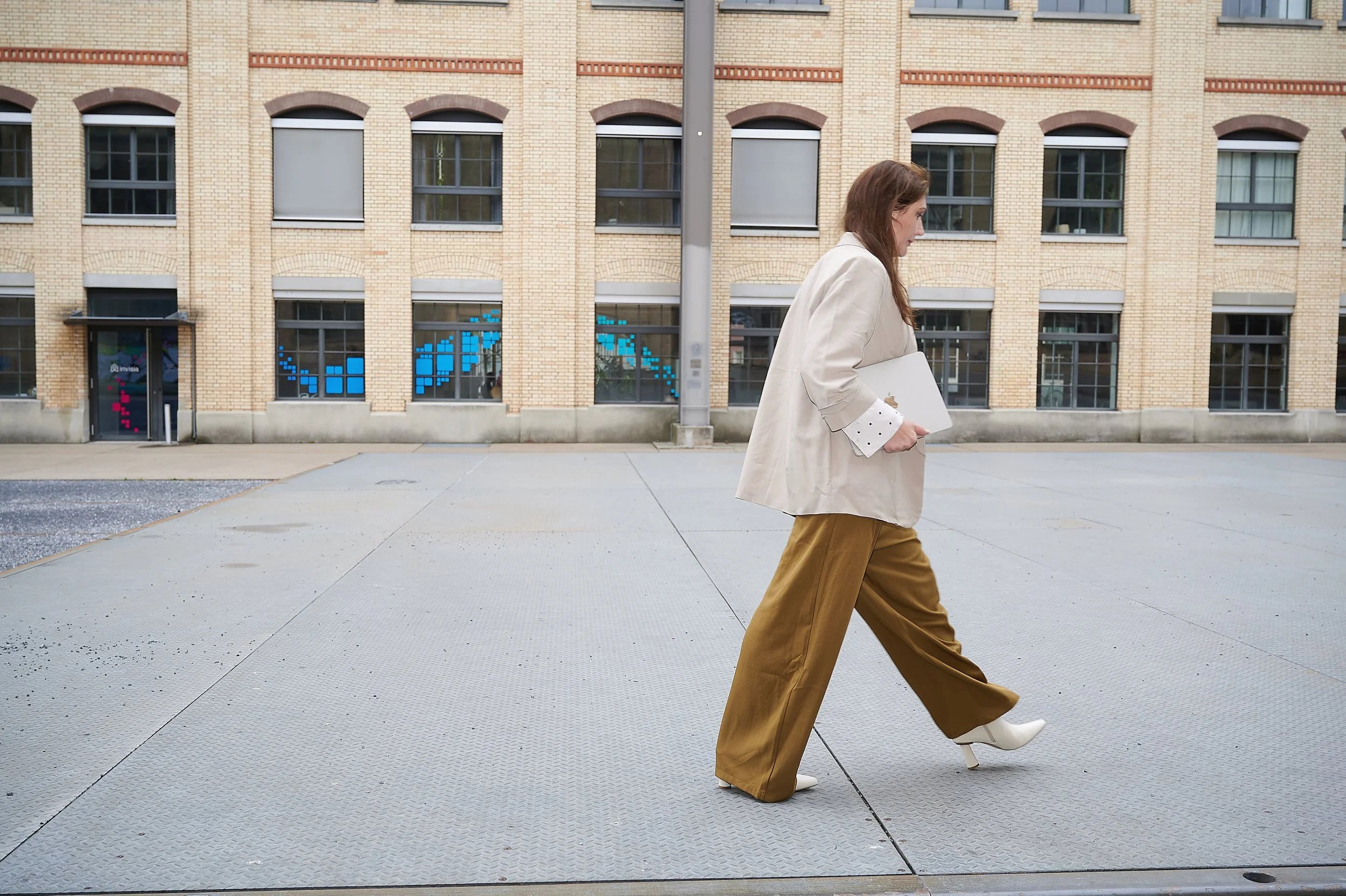 Frau mit braunen Haaren trägt Weiß, beige Mantel, braune Hosen und weiße Stiefel, geht an einem modernen Backstein-Gebäude entlang