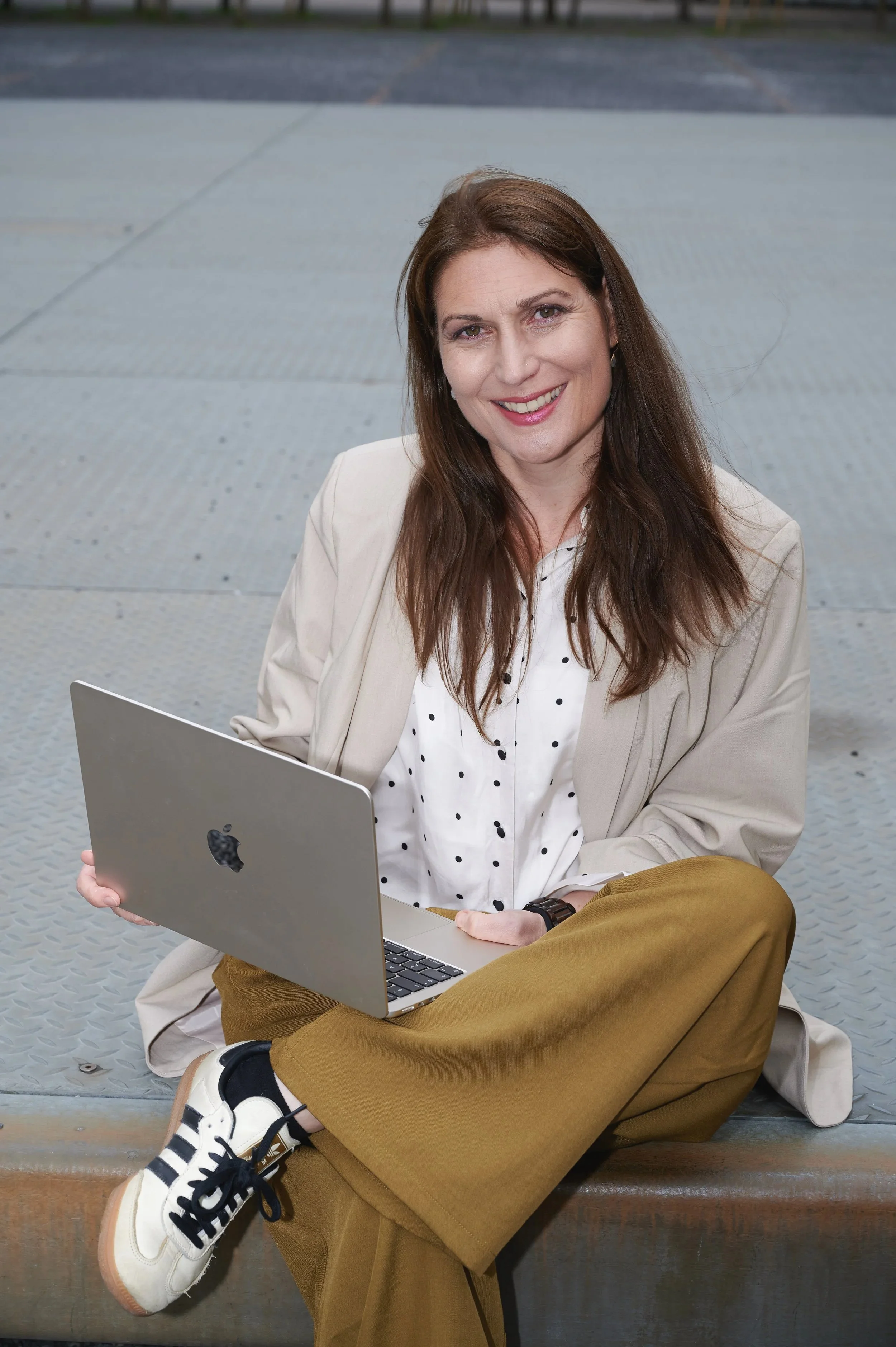 Eine Frau sitzt auf einer Bank im Freien, hält einen Laptop in der Hand und lächelt in die Kamera.