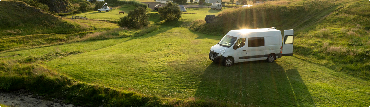 A white van parked on a grassy area in a park with picnic tables and trees, sunlight shining over the scene.