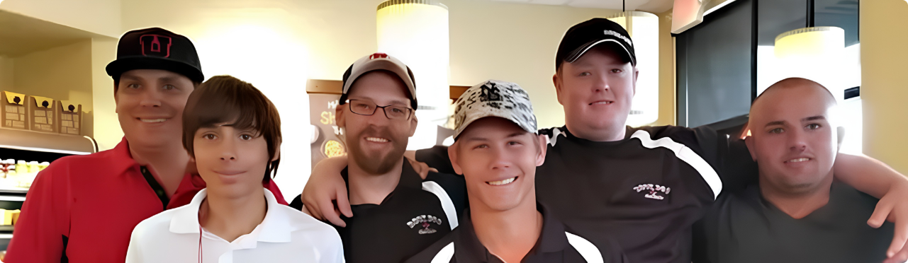 Group of six men and boys smiling, standing close together with arms around each other's shoulders inside a restaurant or cafe.