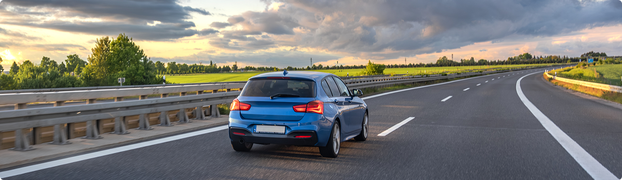A blue hatchback car driving on a highway with guardrails, green trees, and fields nearby during sunset with cloudy skies.