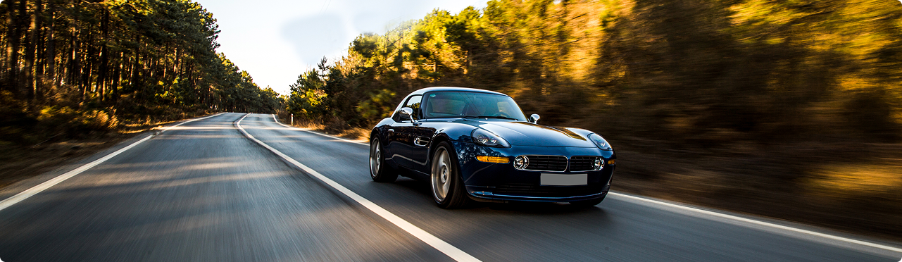 A black convertible sports car driving on a curving road through a wooded area in the countryside with sunlight filtering through trees.
