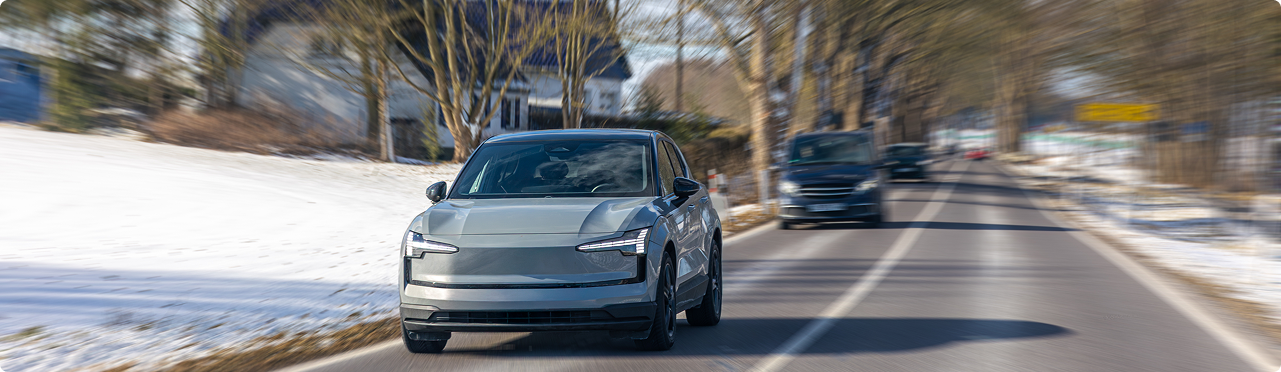 A silver electric vehicle driving on a snowy, rural road with trees and houses in the background.