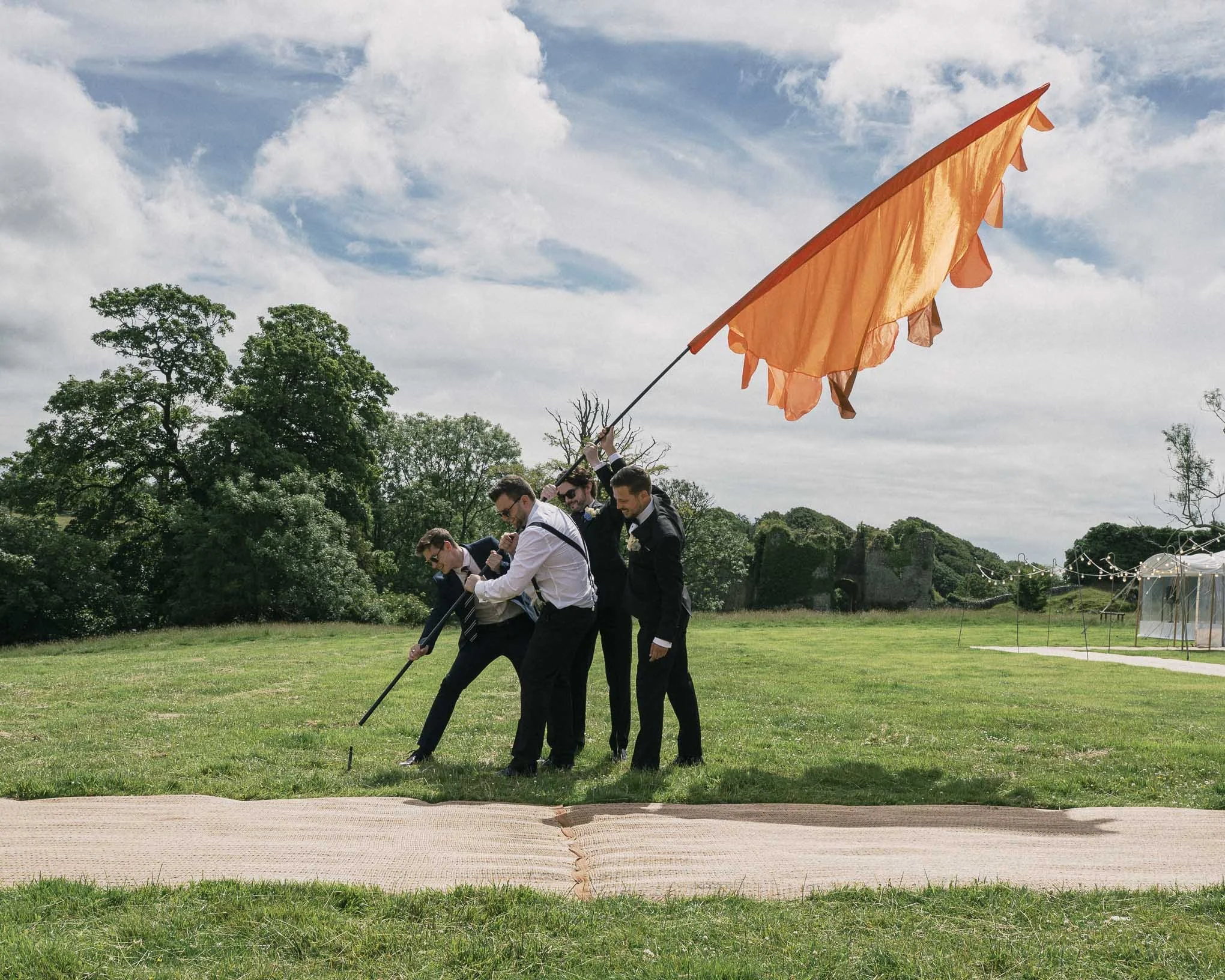 A group of men dressed in suits and ties participating in a golf event, holding an orange flag and walking on a green golf course with trees under a partly cloudy sky.