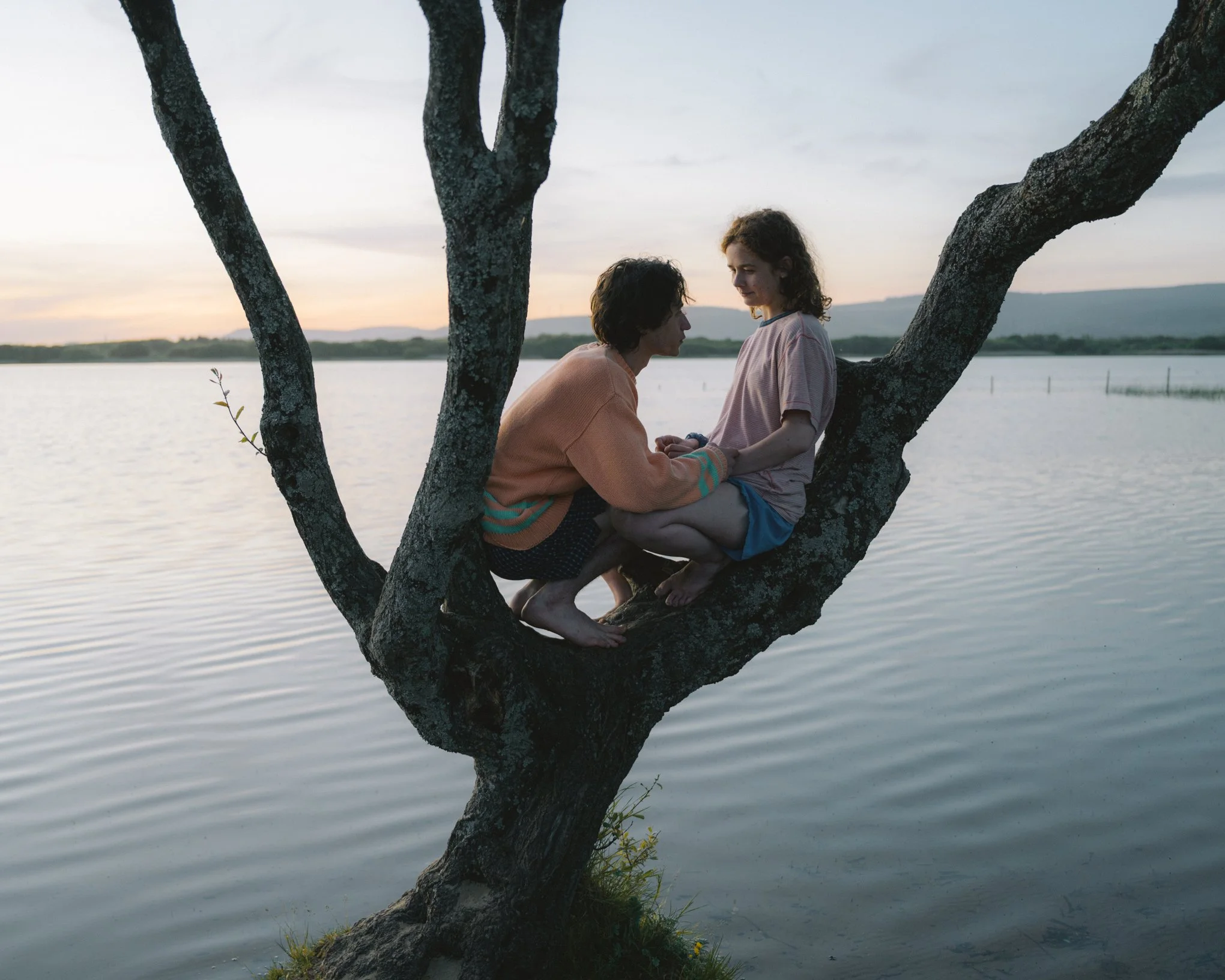 A person and a girl sit on a tree branch over water at sunset, with the person holding the girl's hands and both smiling.