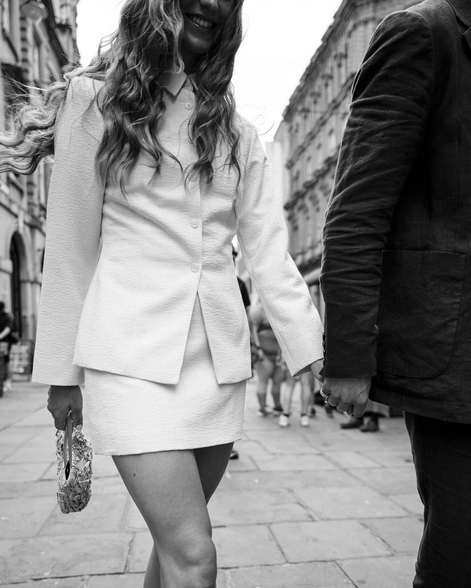A black and white photo of a smiling woman with long, curly hair holding hands with a man on a city street, with buildings in the background.