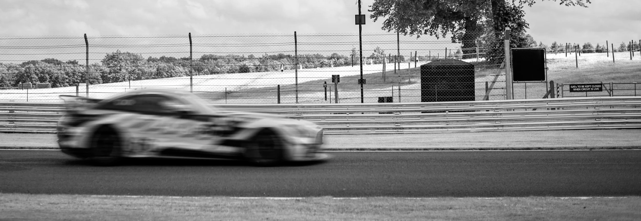A racing car blurred in motion on a racetrack with fences and trees in the background, in black and white.
