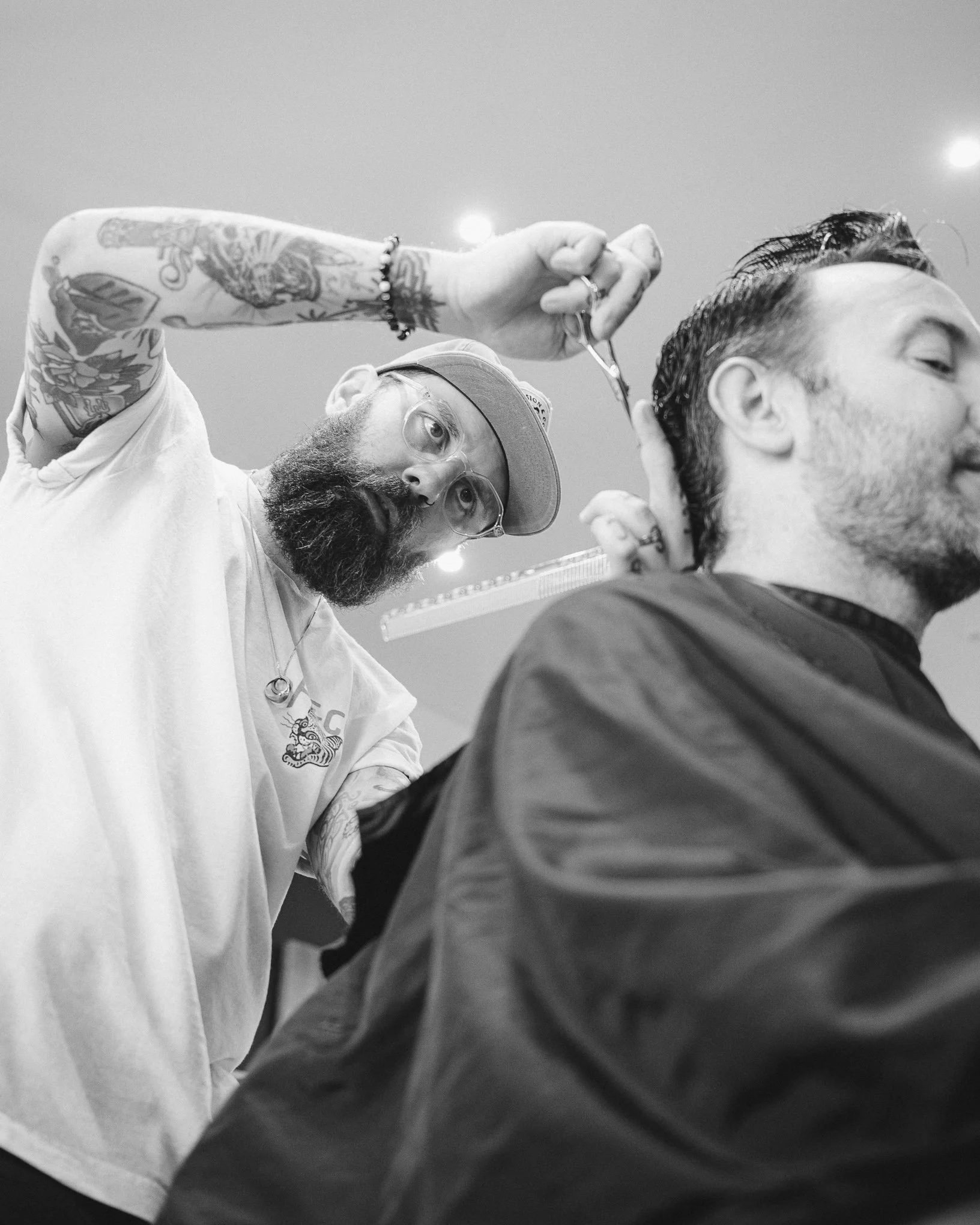 Tattooed man with glasses receiving a haircut from a barber.