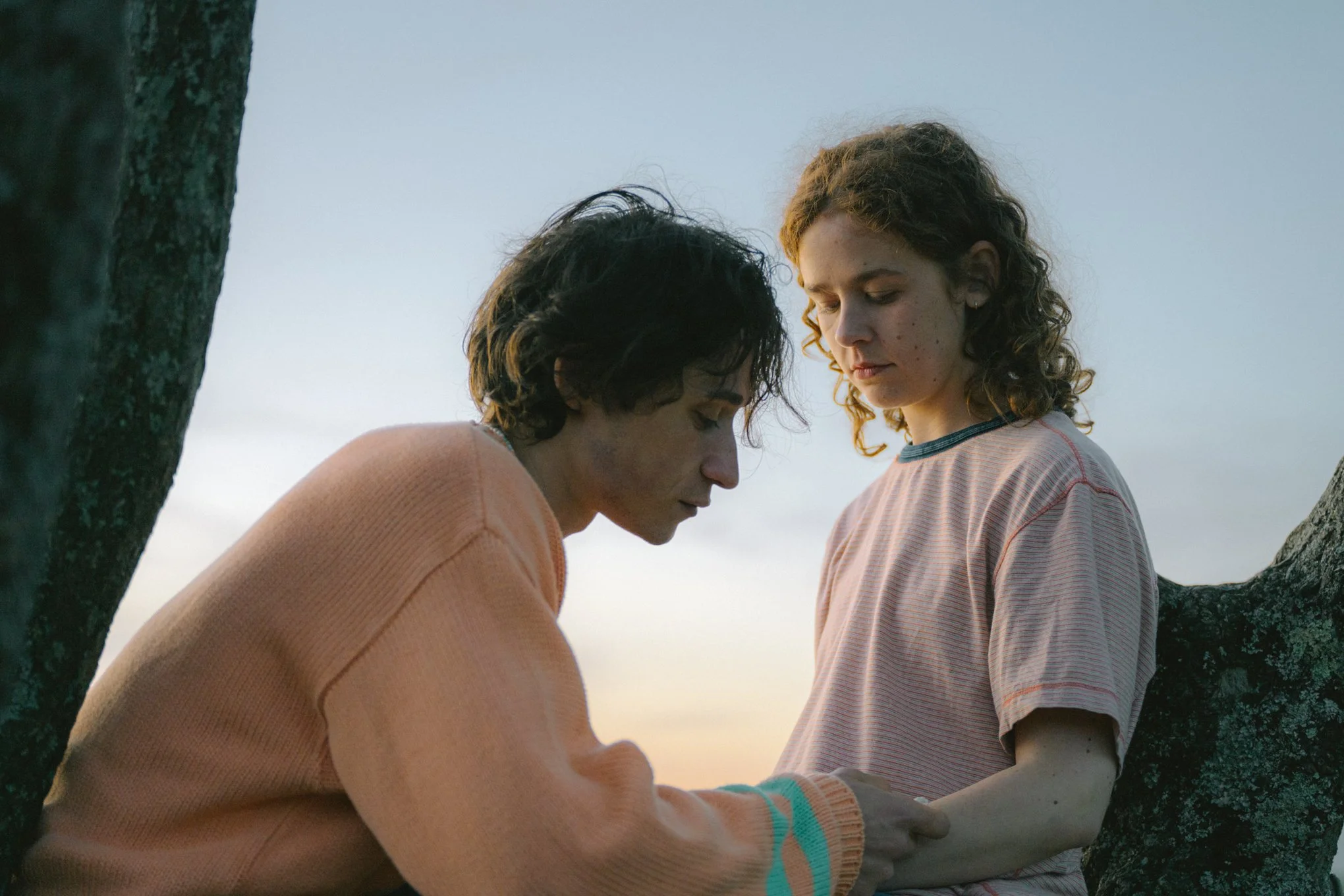 Two young women with curly hair are leaning close to each other outdoors near a tree during sunset, sharing an intimate moment.
