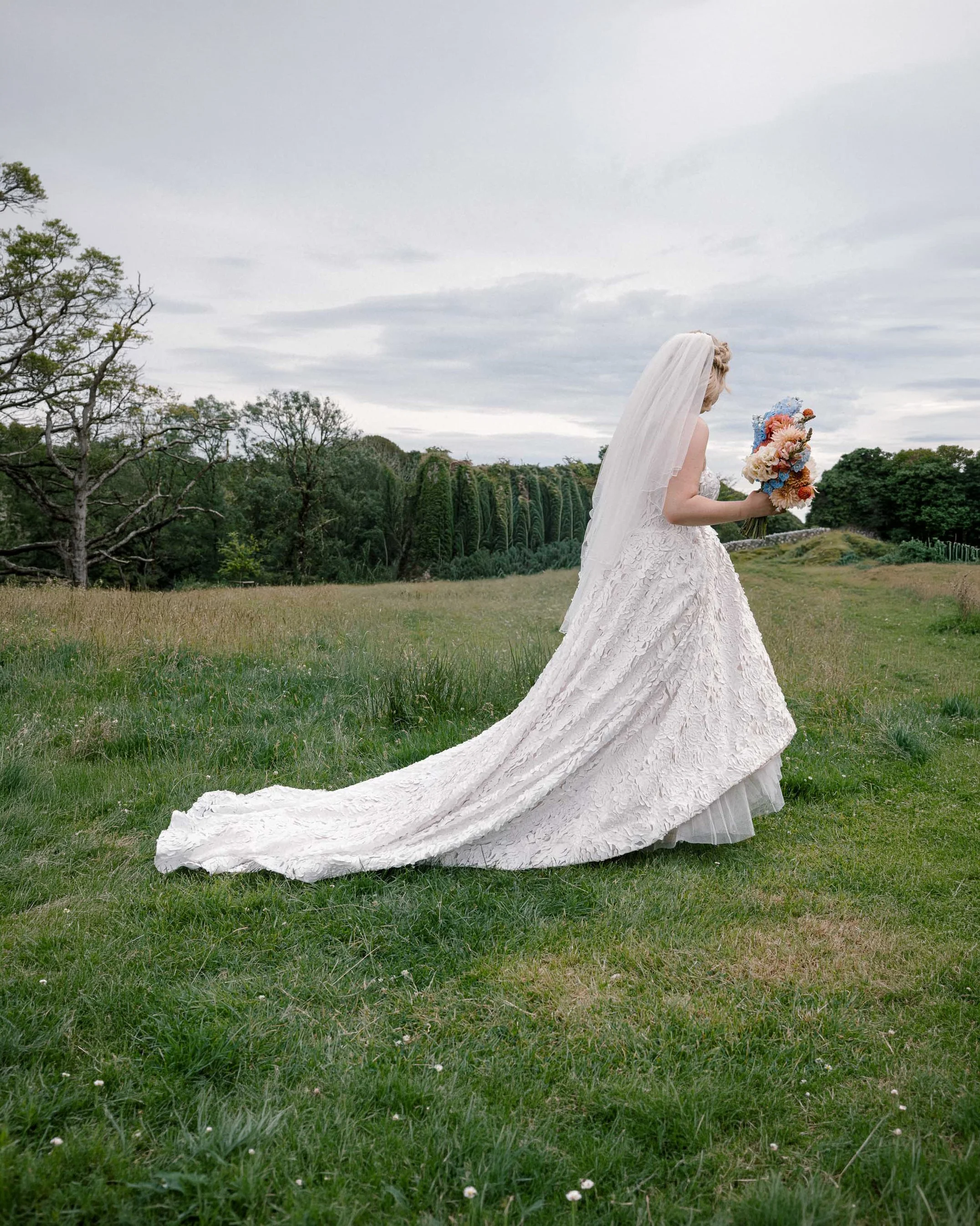 A person in a white wedding dress with a long train and veil standing in a grassy field holding a colorful bouquet, with trees and cloudy sky in the background.