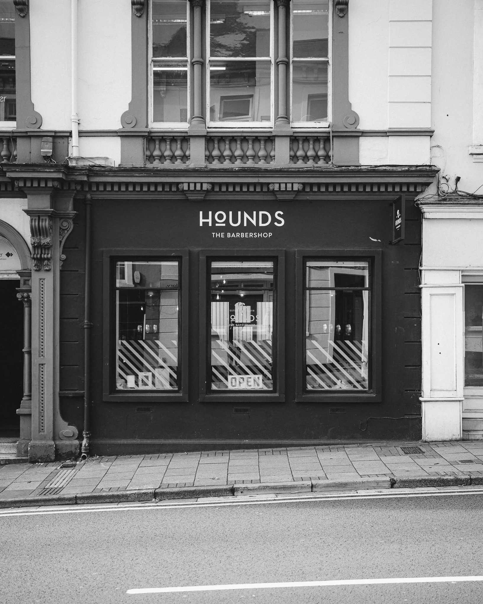 A storefront with a sign that reads 'HOUNDS THE BARBERSHOP' located on a city street sidewalk. The building has ornate architectural details and two large windows.
