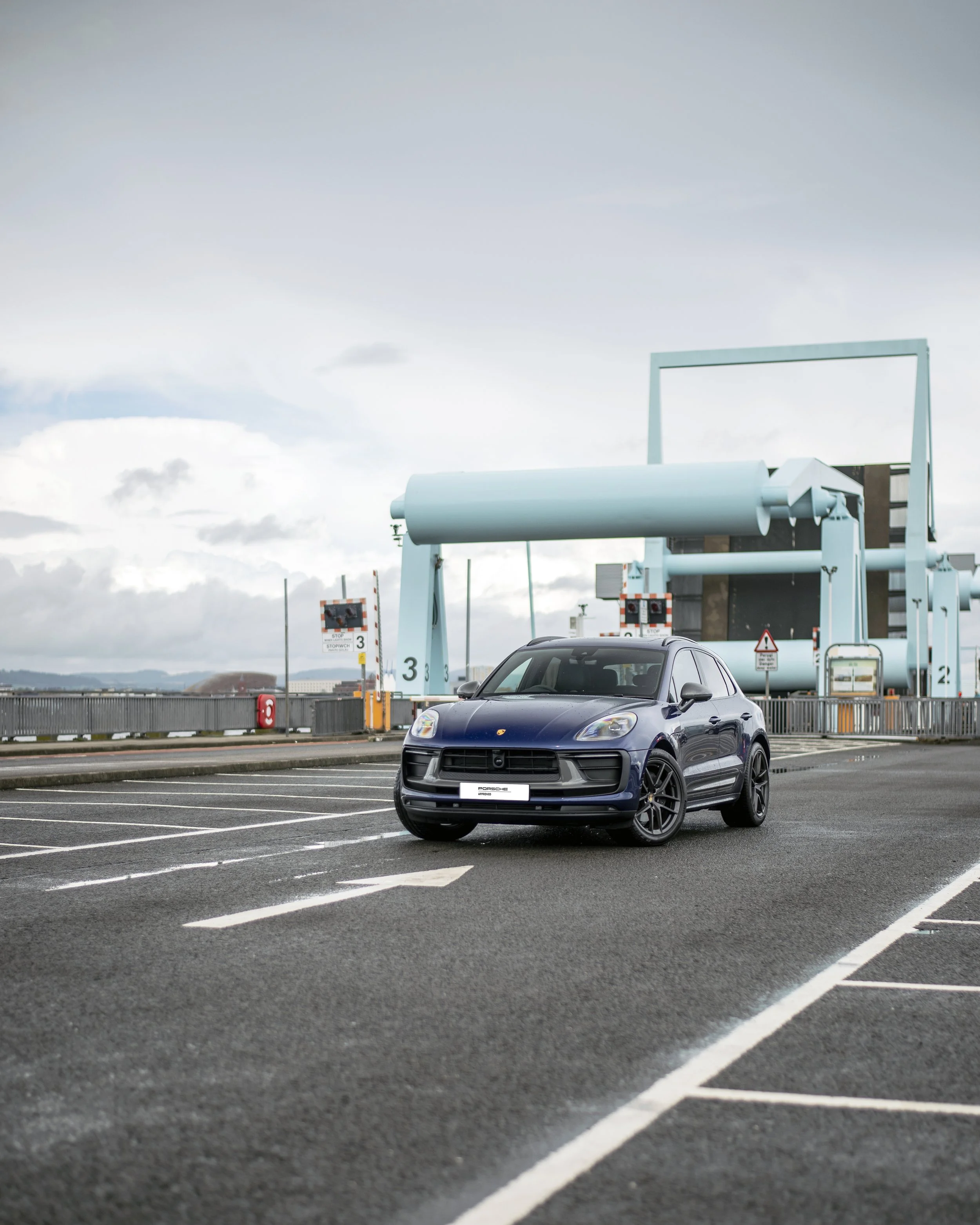A dark blue Porsche Macan SUV parked in an outdoor parking lot with marked spaces, surrounding fencing, and road signs, under a cloudy sky.