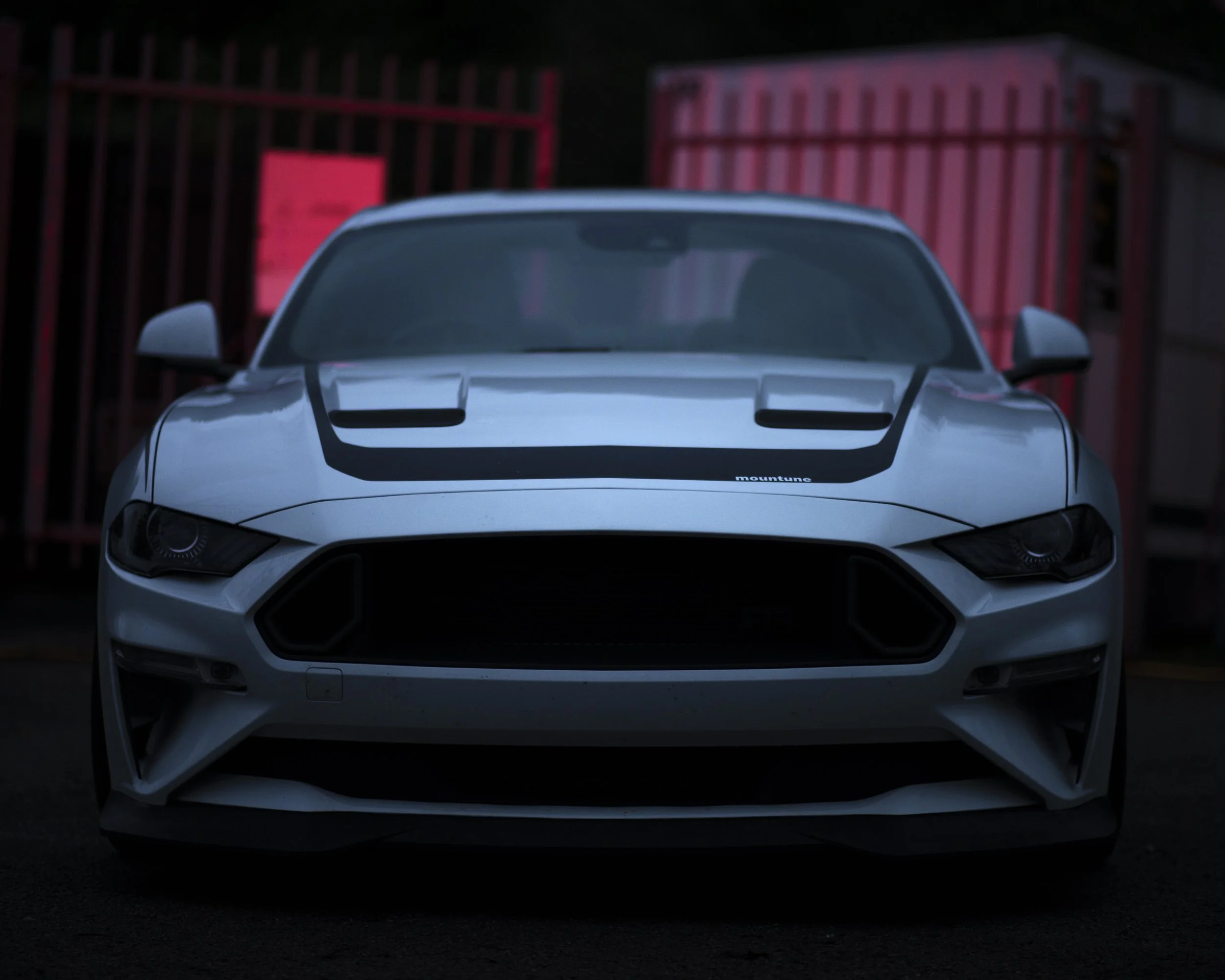 Front view of a white sports car parked outdoors at night, with red fences in the background.