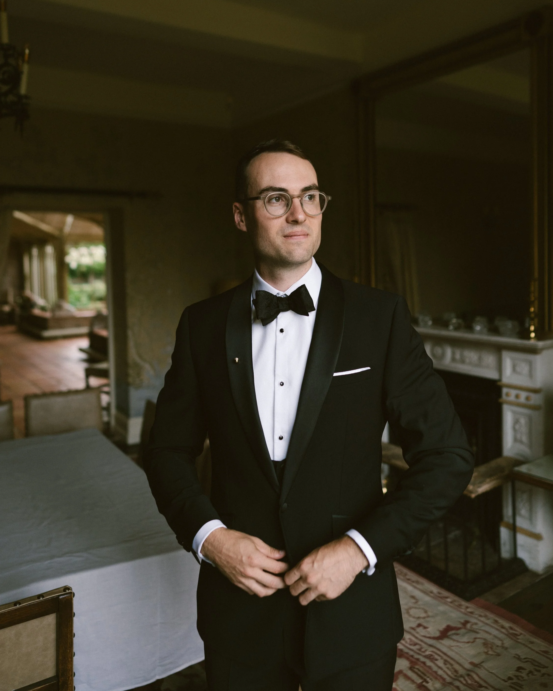 A man in a black tuxedo with a bowtie and glasses standing indoors in a dimly lit room.