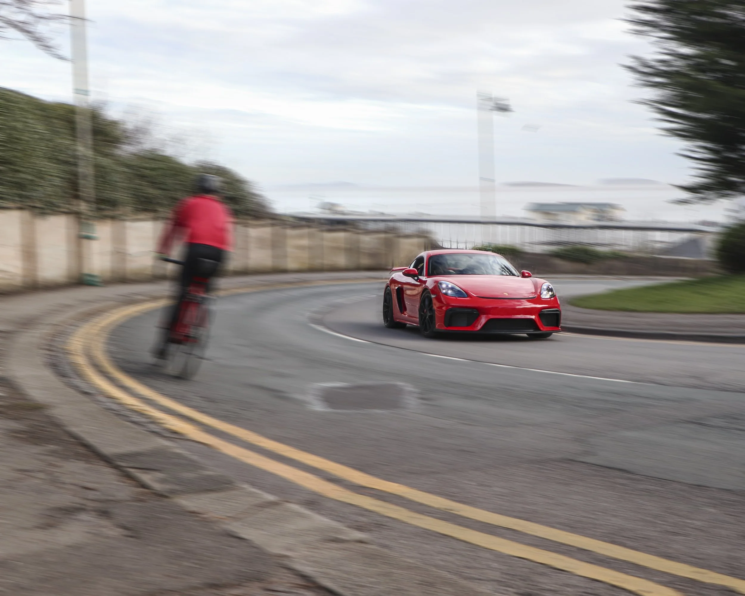 A red sports car driving on a curved street with a cyclist in a red jacket and black helmet riding beside it. There are trees and a cloudy sky in the background.