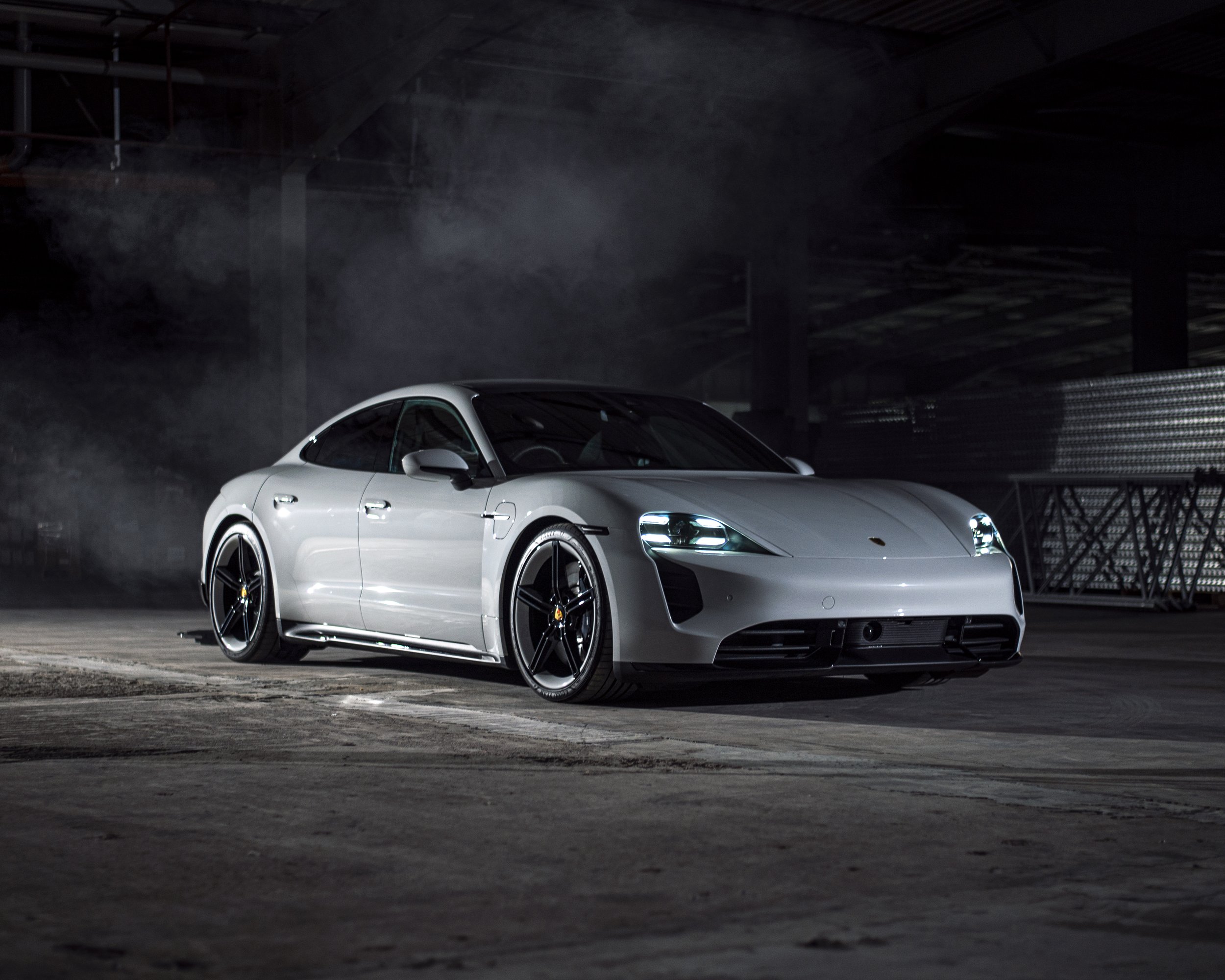 A silver sports car with black wheels parked inside a dark warehouse or industrial space, with visible smoke or mist around the vehicle.