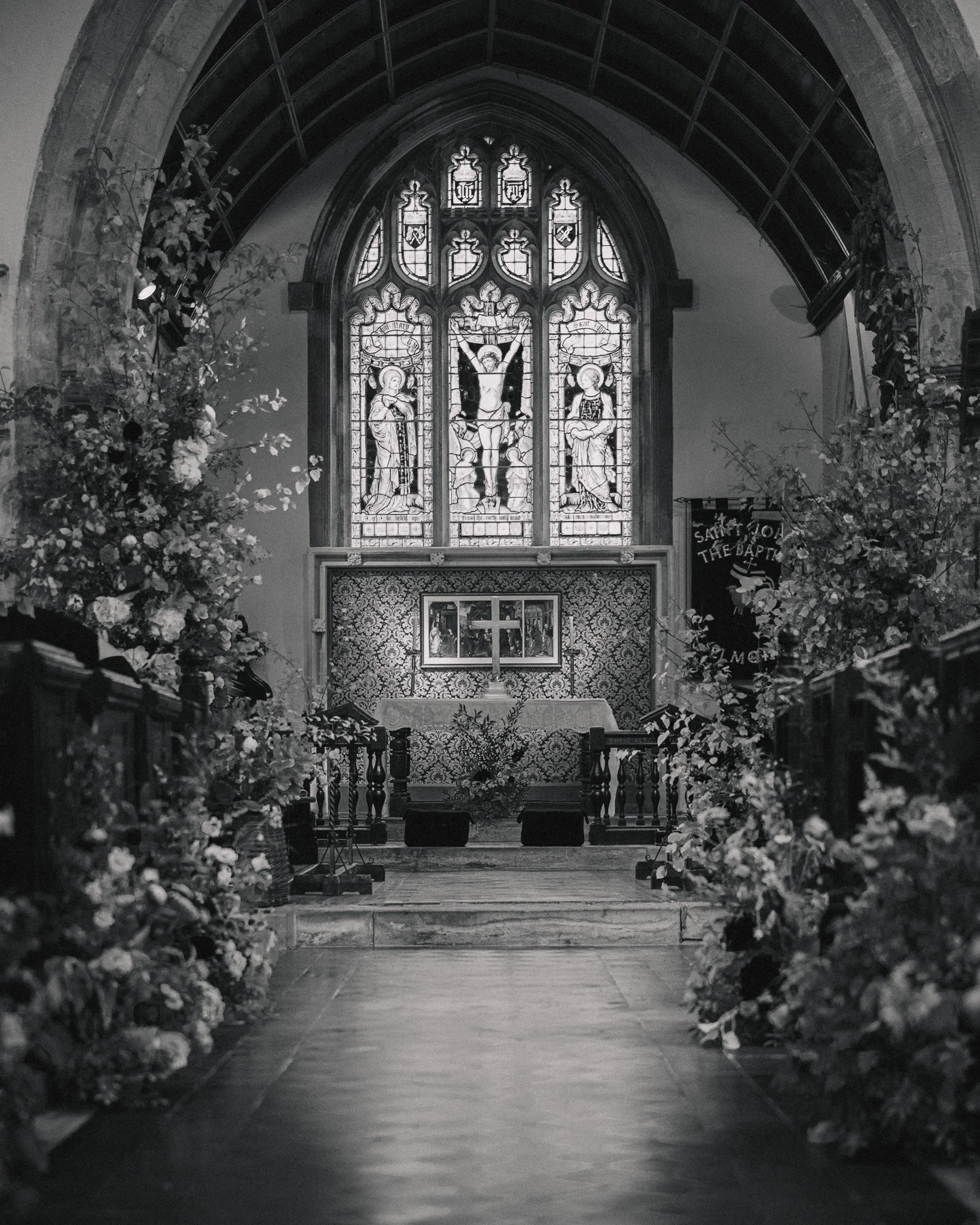 Inside a church, an altar with flowers on both sides leads up to a stained glass window depicting religious figures, including Jesus on the cross, flanked by two saints, with a decorative arched design.