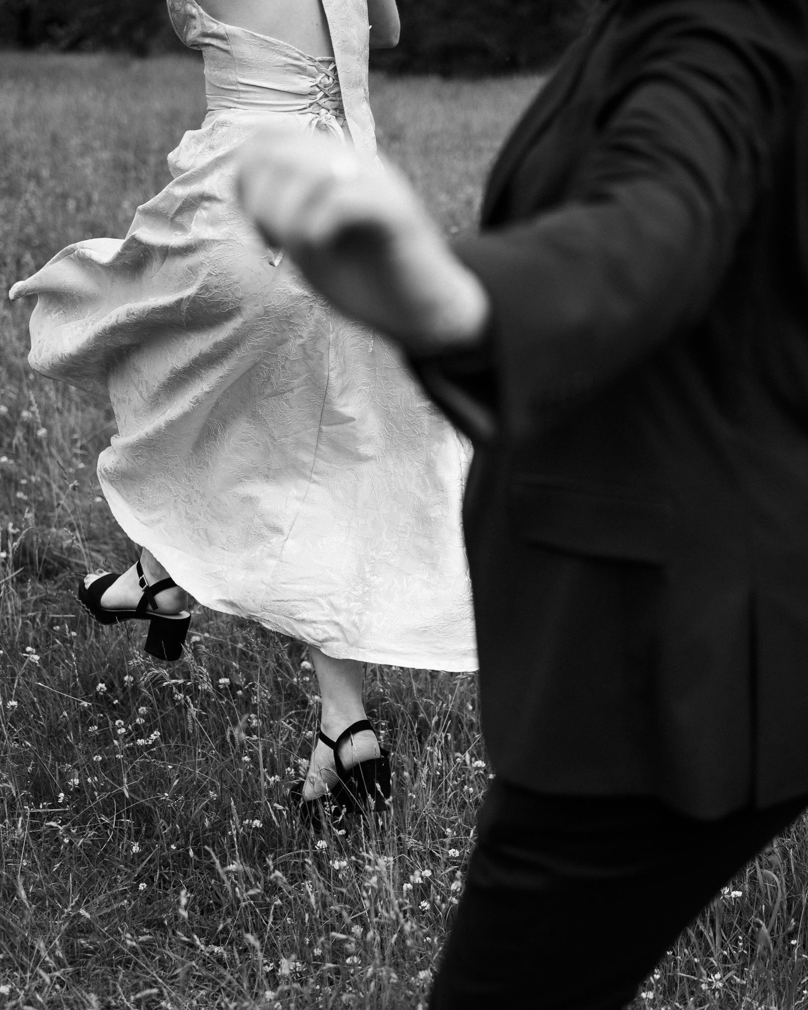 A black and white photo of a woman in a light dress holding hands with a man dressed in dark clothing walking through a grassy field.