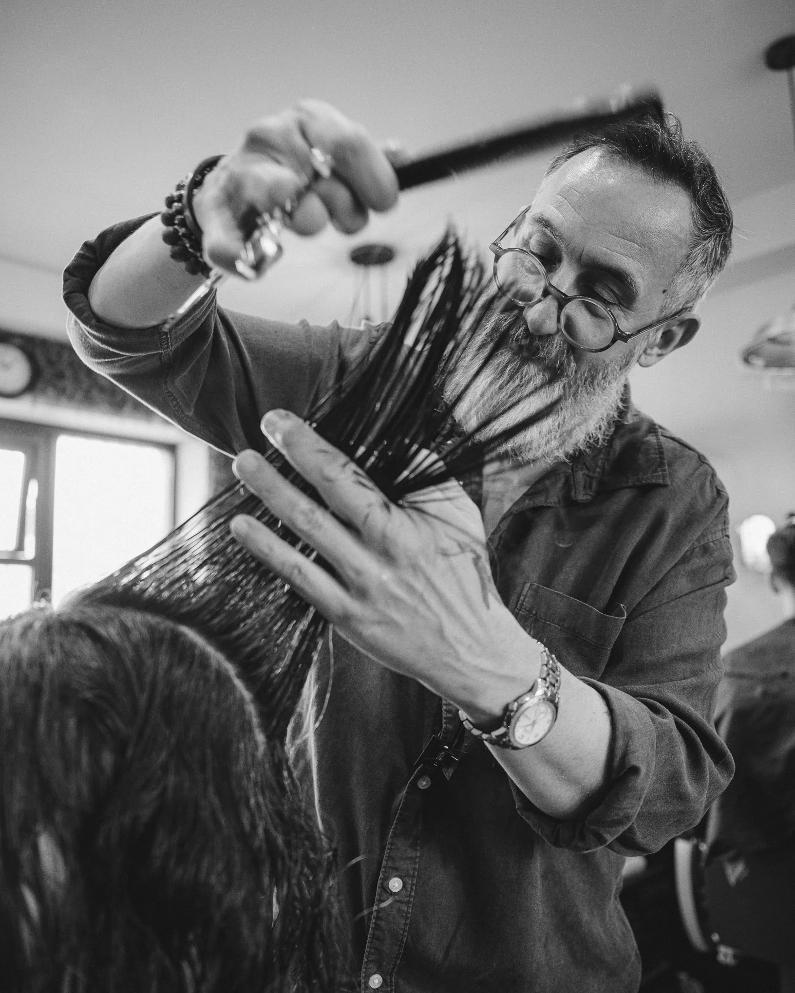 A hairstylist with gray hair, glasses, and wearing rings and bracelets, is cutting a client's hair using scissors and a comb in a salon.