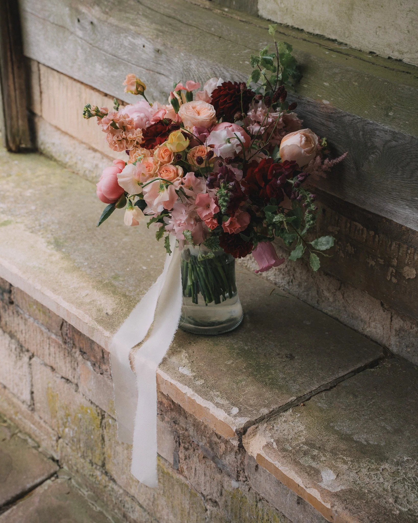 A bouquet of pink, red, and peach flowers with green foliage in a clear glass jar, placed on a weathered brick surface against a wooden wall.