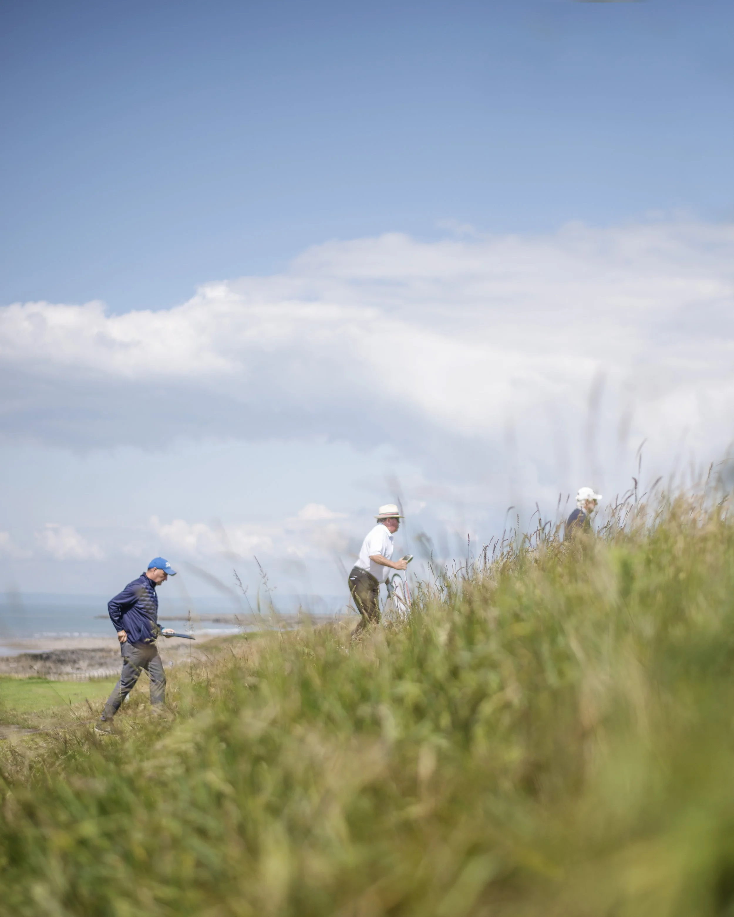 People walking along a grassy beach area under a blue sky with scattered clouds.