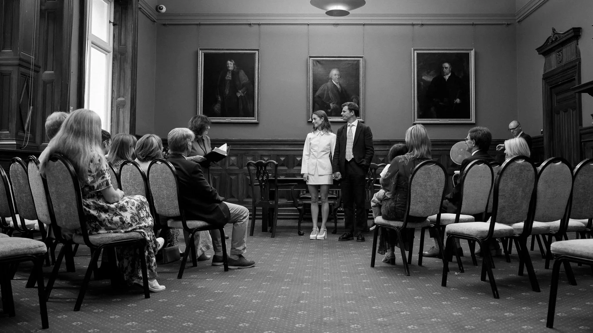 A black-and-white photo of a formal room with a group of people attending a ceremony or event. In the center, a man in a suit and a woman in a white dress are holding hands and facing a woman with a book, possibly a judge or officiant. The room has framed portraits of historical figures on the wall, and several seated guests are watching the scene.