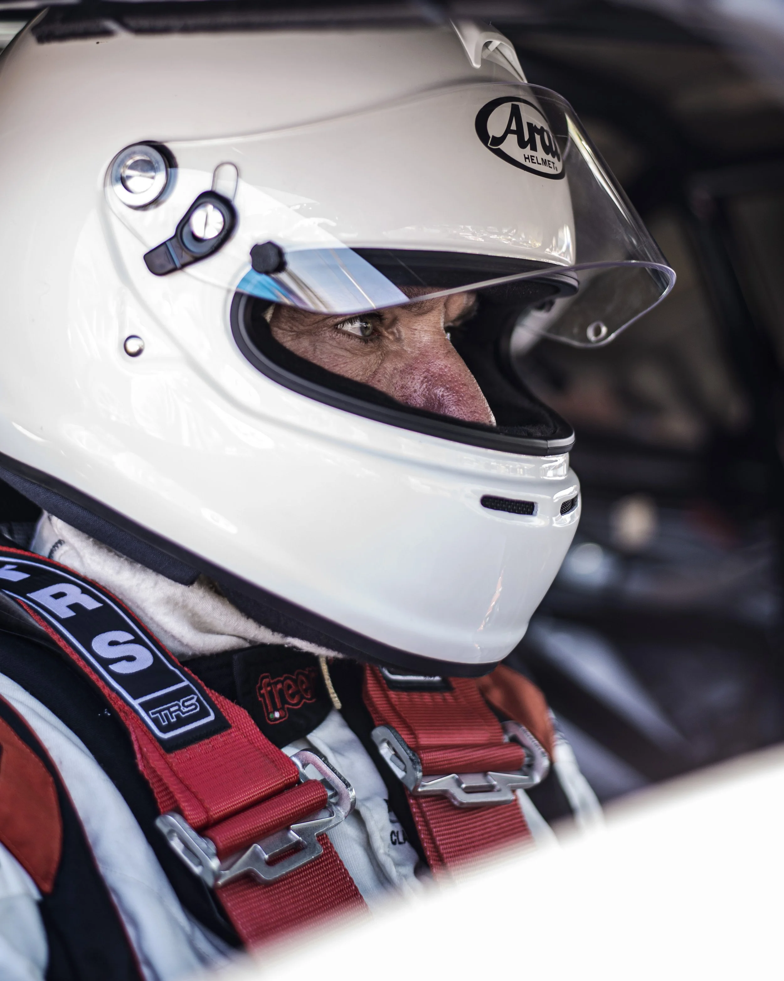 A race car driver wearing a white helmet with a clear visor, looking focused. The helmet has a logo that reads 'Arai Helmet.' The driver is dressed in a racing suit with red and black harness straps.