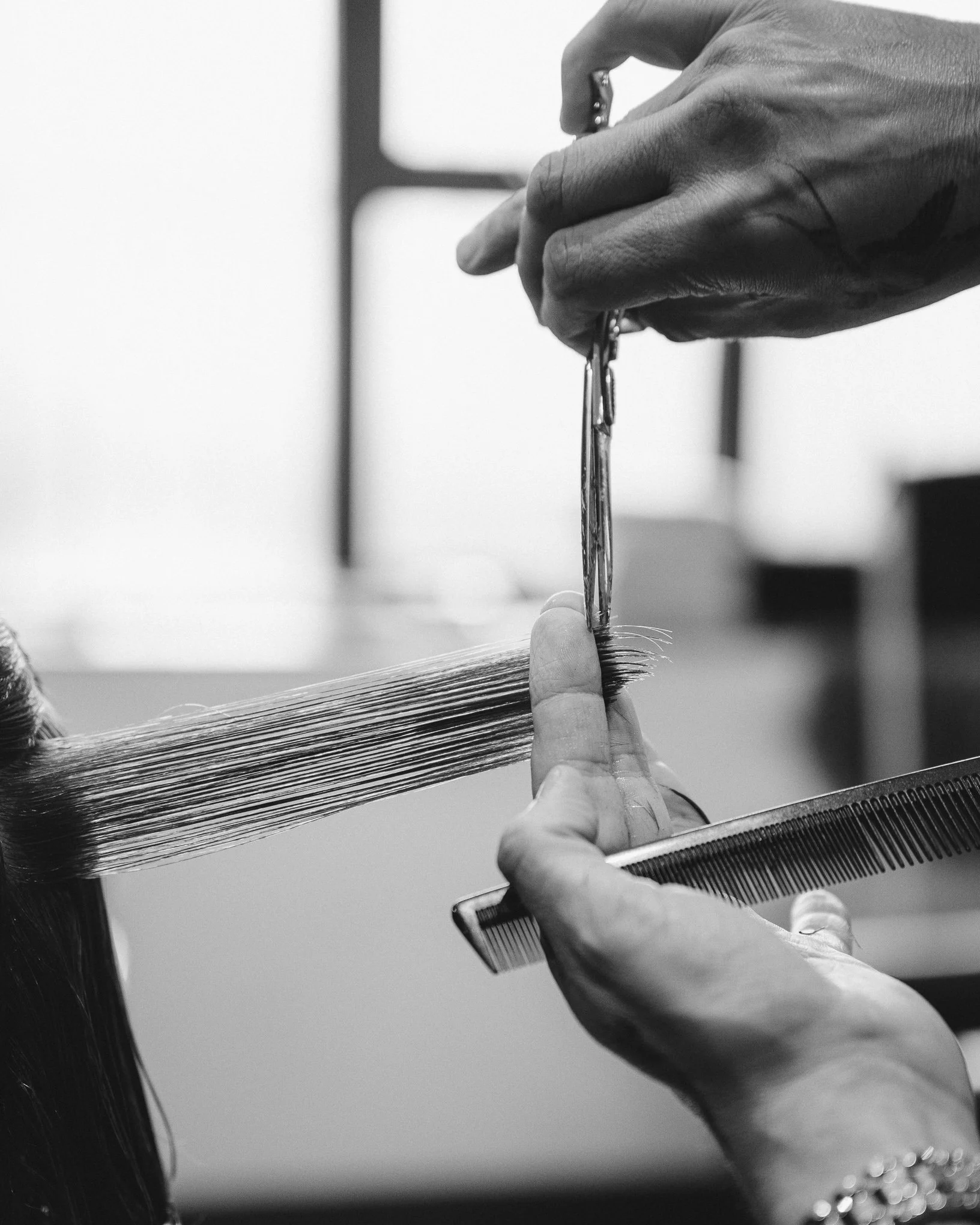 A person with jewelry on their wrist is combing or braiding a long, dark strand of hair with a small metal tool.