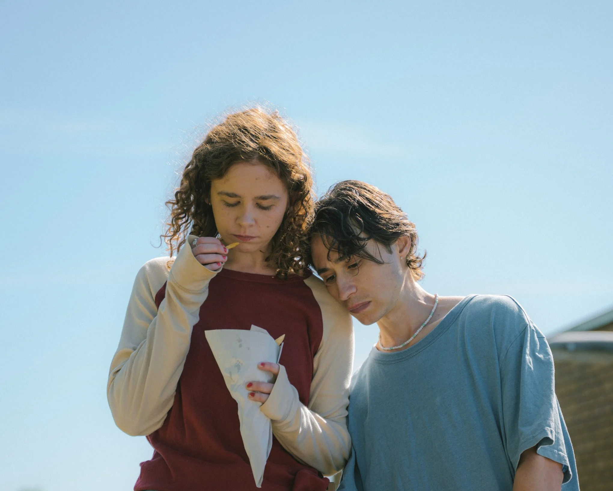 Two teenagers, a girl with curly brown hair and a boy with short dark hair, are looking at a piece of paper or card. The girl is holding it with both hands and appears to be reading or inspecting it. They are outdoors under a clear blue sky.