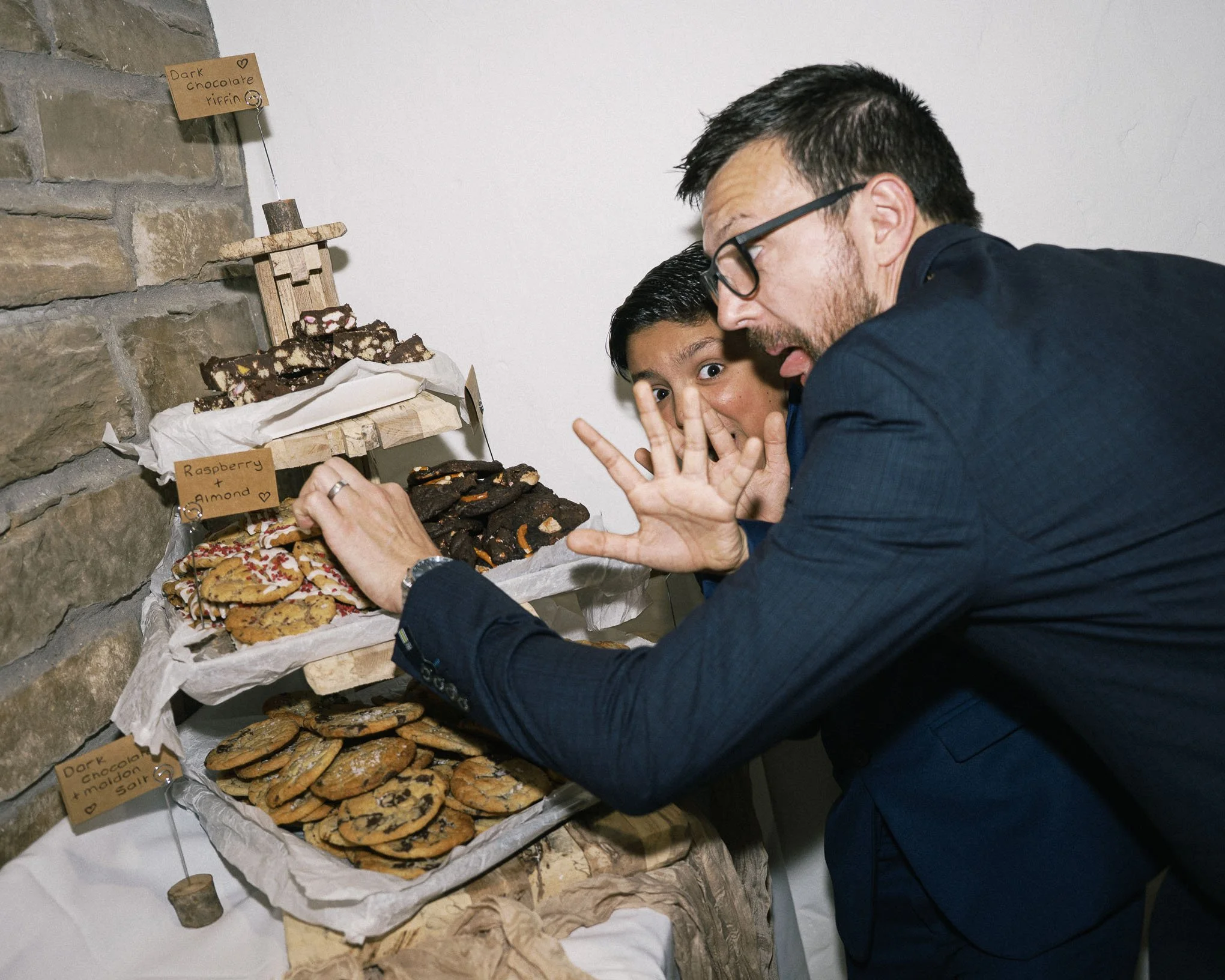A man and a young boy at a dessert table with various cookies and snack bars, some labeled with handwritten tags such as "Dark chocolate + almond salt" and "Raspberry + almond." The man is making a playful face with his tongue out, while the boy shows a finger gesture and wide-eyed expression.