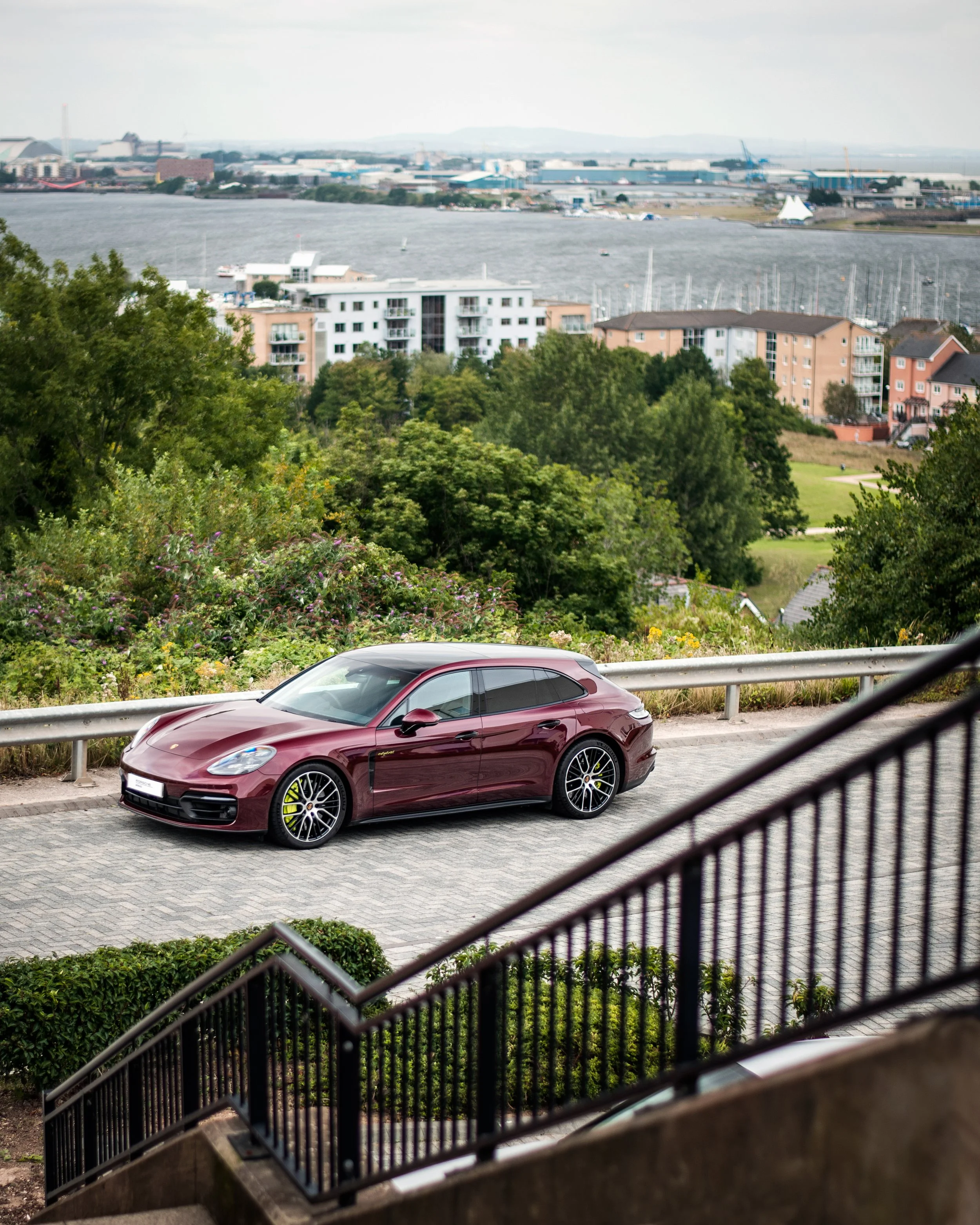 A view of a cityscape with a maroon sports car parked on a paved area in the foreground. The scene is viewed from a staircase with a black metal railing, overlooking green trees, residential buildings, and a distant industrial area across a body of w