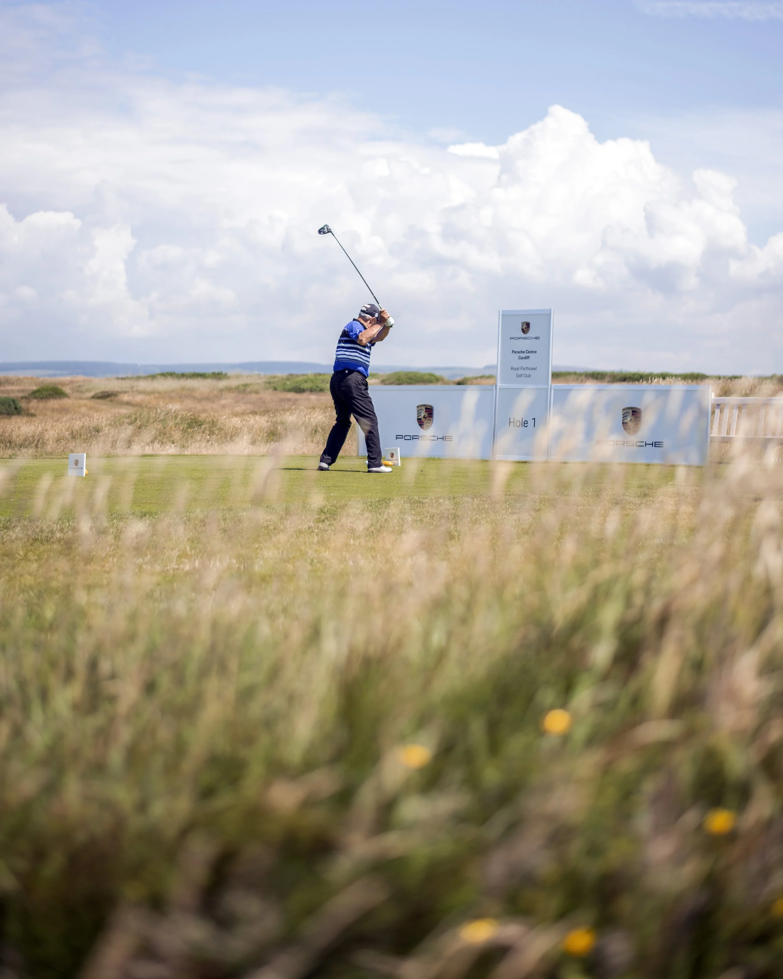 A person playing golf on the driving range at a golf course, with open fields and a partly cloudy sky in the background.