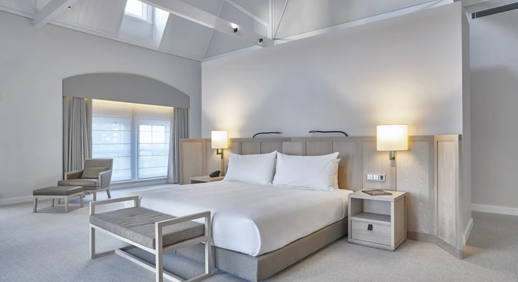 A modern, minimalist hotel bedroom with a large bed, two side tables with lamps, a sitting chair and ottoman near the window, and skylights in the ceiling.