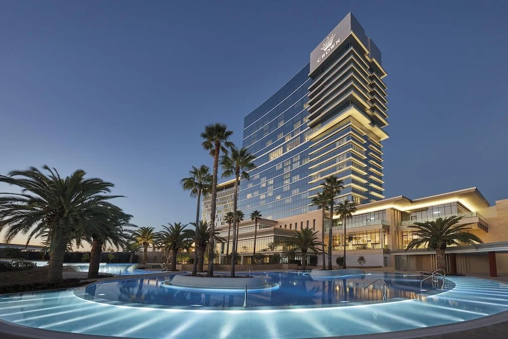 A modern high-rise hotel with palm trees and a lit pool in the foreground at dusk.
