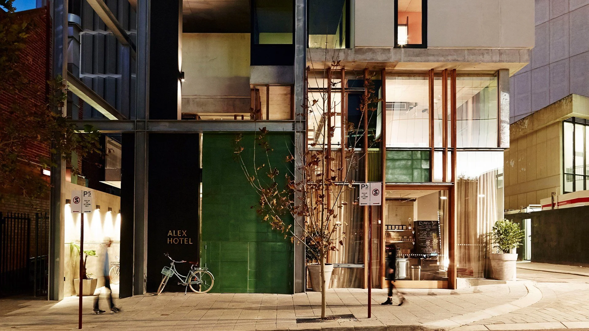 Nighttime view of the exterior of a modern building with large glass windows, a green tiled section with the sign 'Alex Hotel,' and a small tree in front. Pedestrians and a bicycle are visible on the sidewalk.