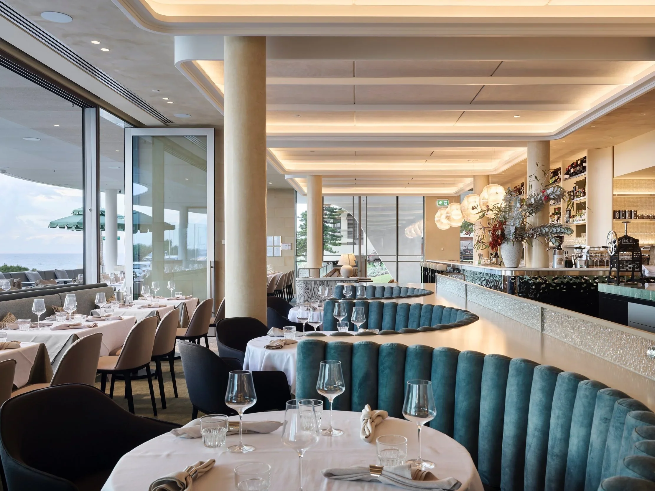 Elegant restaurant interior with large windows, white tablecloths, wine glasses, modern chairs, and decorative lighting, overlooking a view outside with umbrellas and trees.
