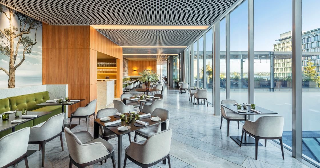 Modern restaurant with large floor-to-ceiling windows, wooden accents, and a mix of green and white seating, set for dining.