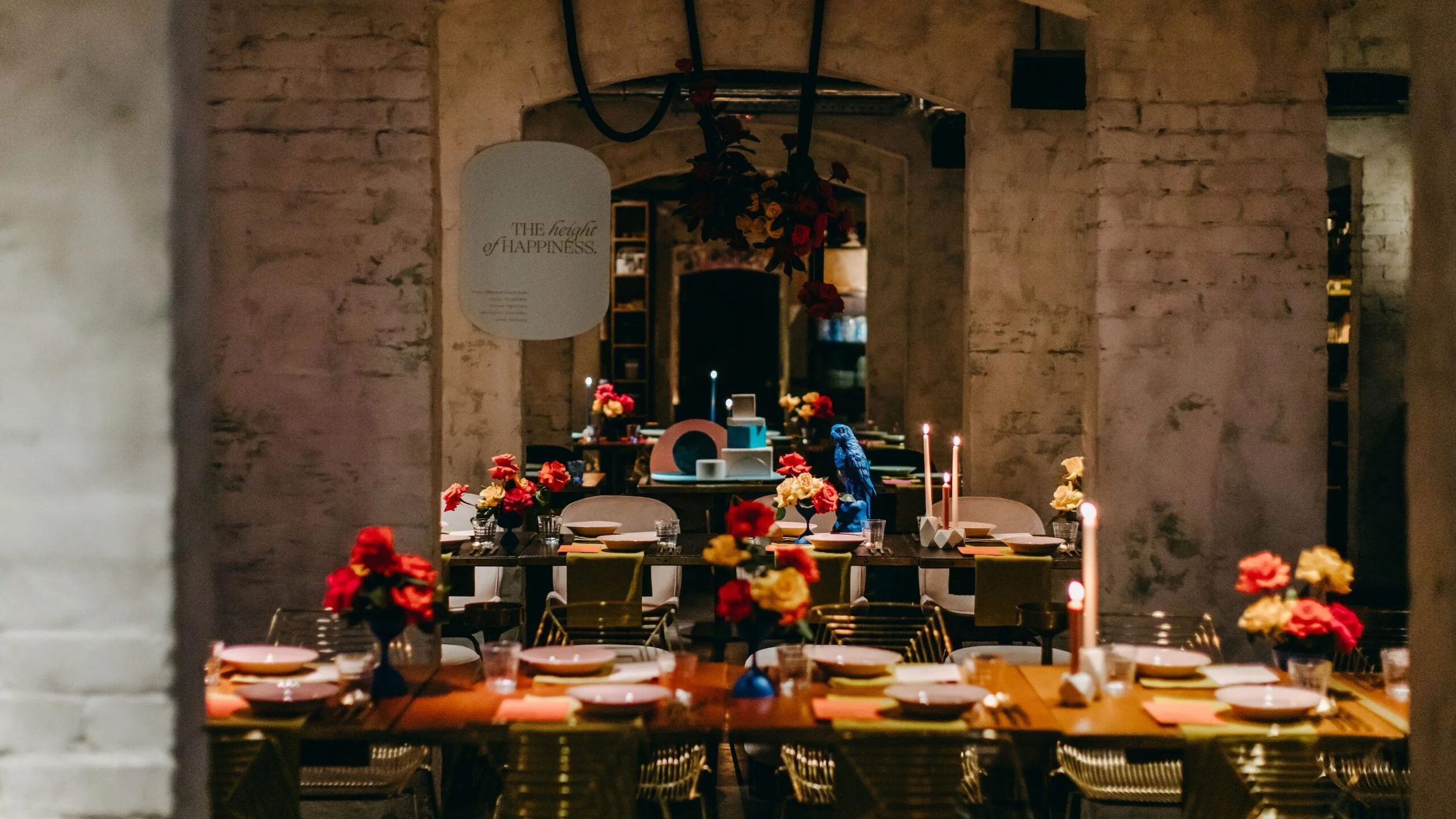 A cozy restaurant or event space decorated with vibrant red, pink, and yellow flowers, candles, and modern tableware. The background features a rustic brick wall and a sign that says 'The height of happiness.'