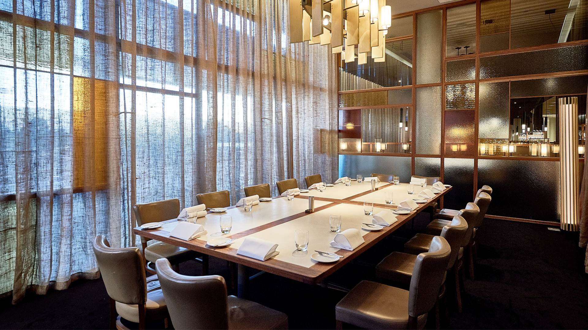 A restaurant dining area with a large rectangular table set with white napkins, glasses, and silverware. The room features floor-to-ceiling window curtains, modern lighting fixtures, and a partition wall with glass and textured panels.