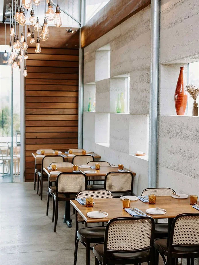 Interior view of a modern restaurant with wooden and concrete walls, hanging Edison-style light bulbs, and tables set with plates, napkins, and amber-colored glasses.