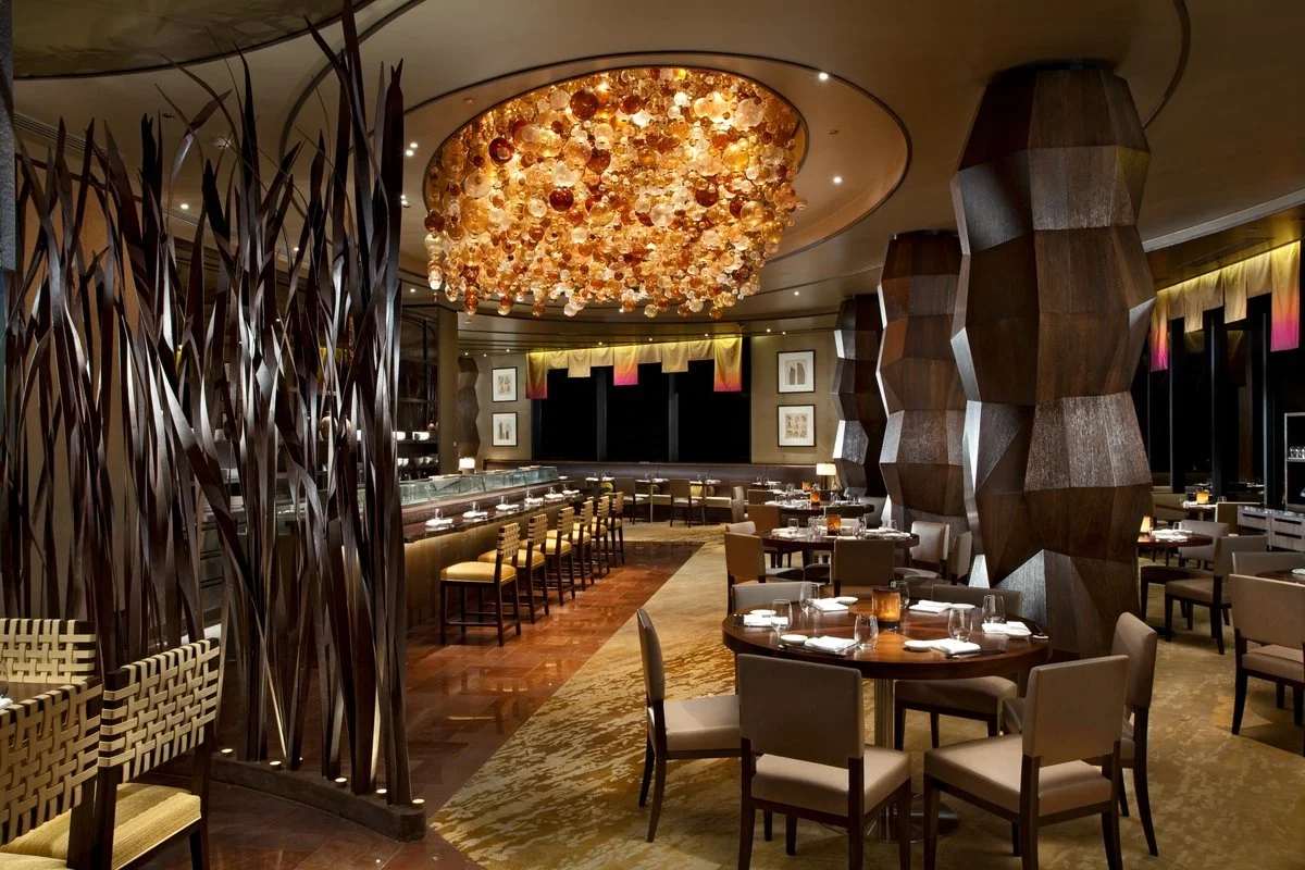 Elegant restaurant interior with modern decor, round dining tables set with plates and glasses, plush chairs, a large circular ceiling fixture with hanging glass orbs, decorative wall art, and intricate wooden pillar designs.