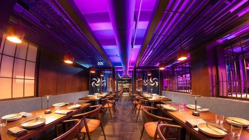 Upscale restaurant with wooden tables set for dining, modern lighting, purple and warm ambient lights, and a contemporary interior design.