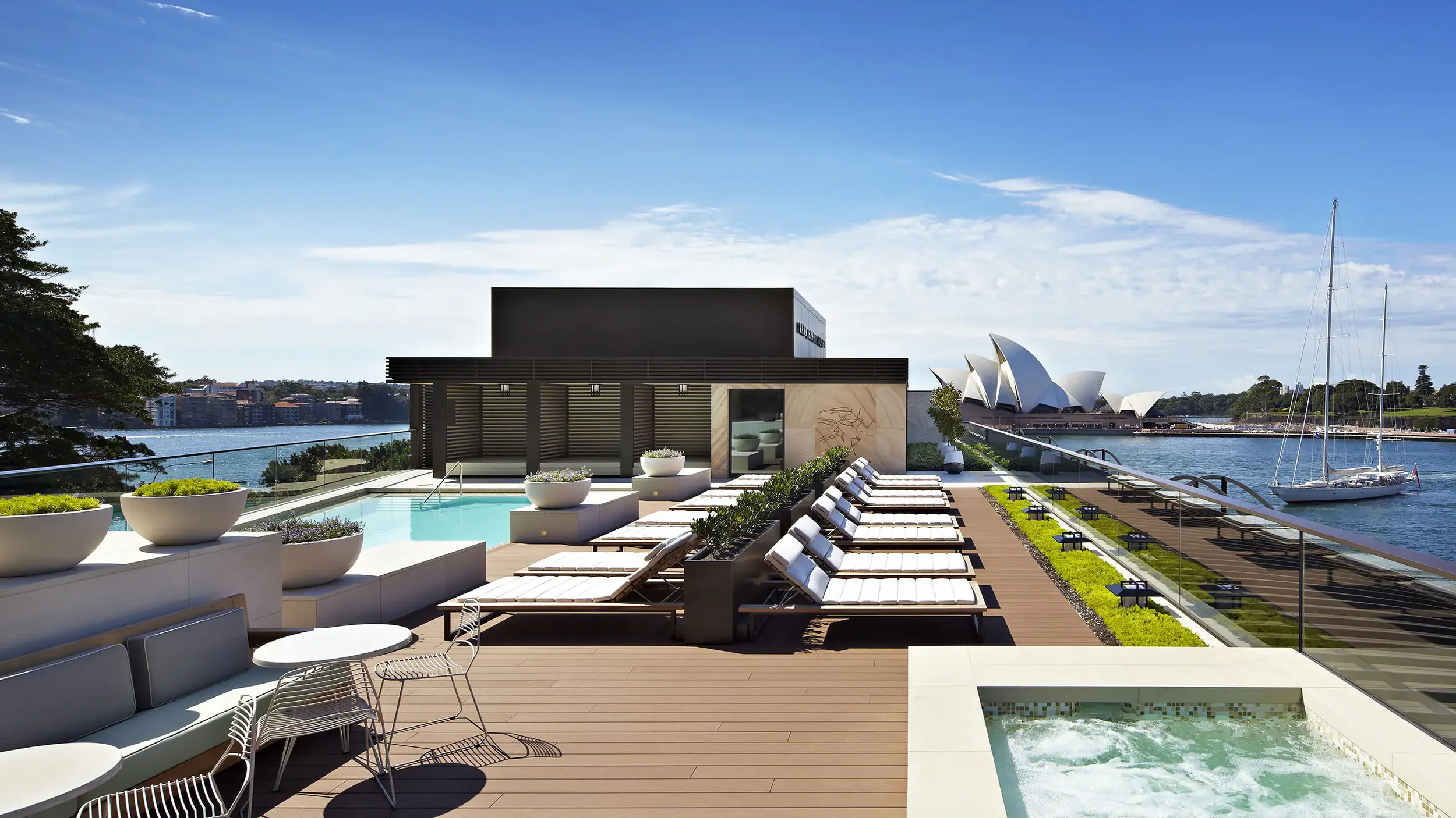 Luxury rooftop pool area with lounge chairs, potted plants, and views of Sydney Opera House and harbor with sailboat under blue sky.
