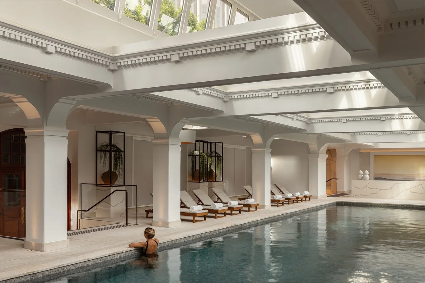Indoor swimming pool with white architectural details and a woman relaxing at the edge of the pool.
