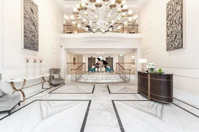 Elegant hotel lobby with white marble floors, a large modern chandelier, and artwork on the walls. There are benches, a dark wood reception desk, and a person walking in the background.