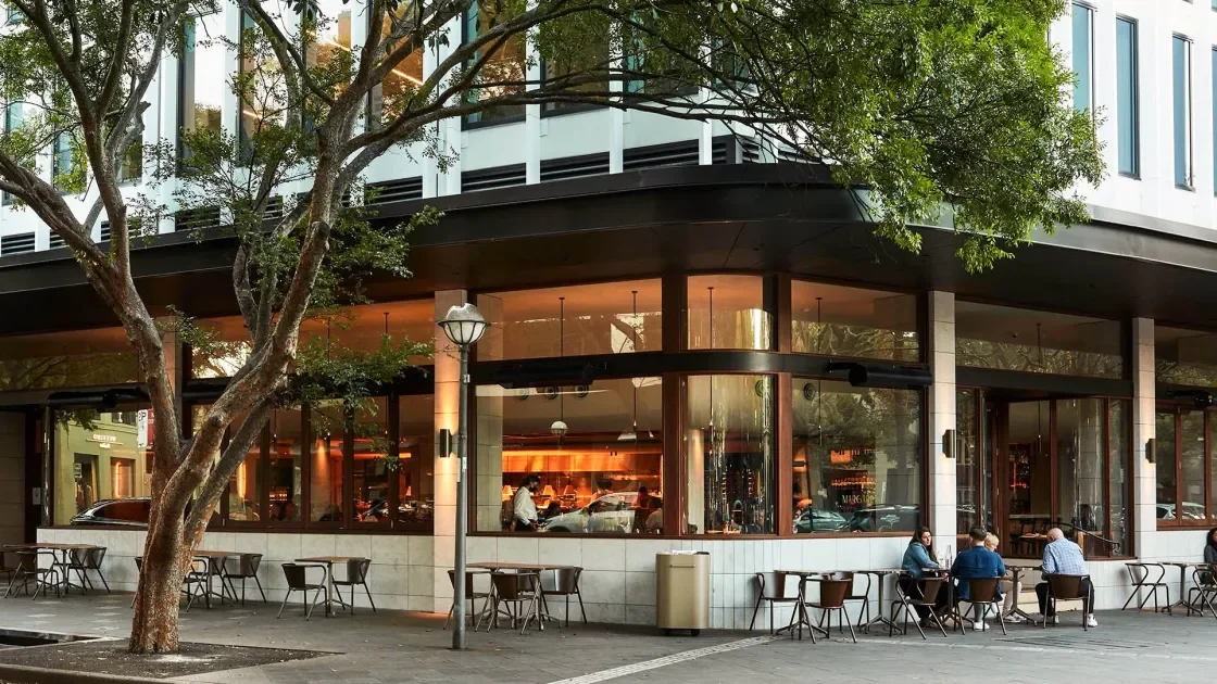 Outside of a modern cafe with large windows, outdoor seating, and a few people sitting at tables.
